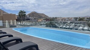 Una piscina en la azotea con sillones reclinables, un puerto deportivo al fondo y un paisaje montañoso.