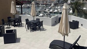 Terraza en la azotea con sillones, mesas y sombrillas, con vistas a un puerto deportivo lleno de barcos.