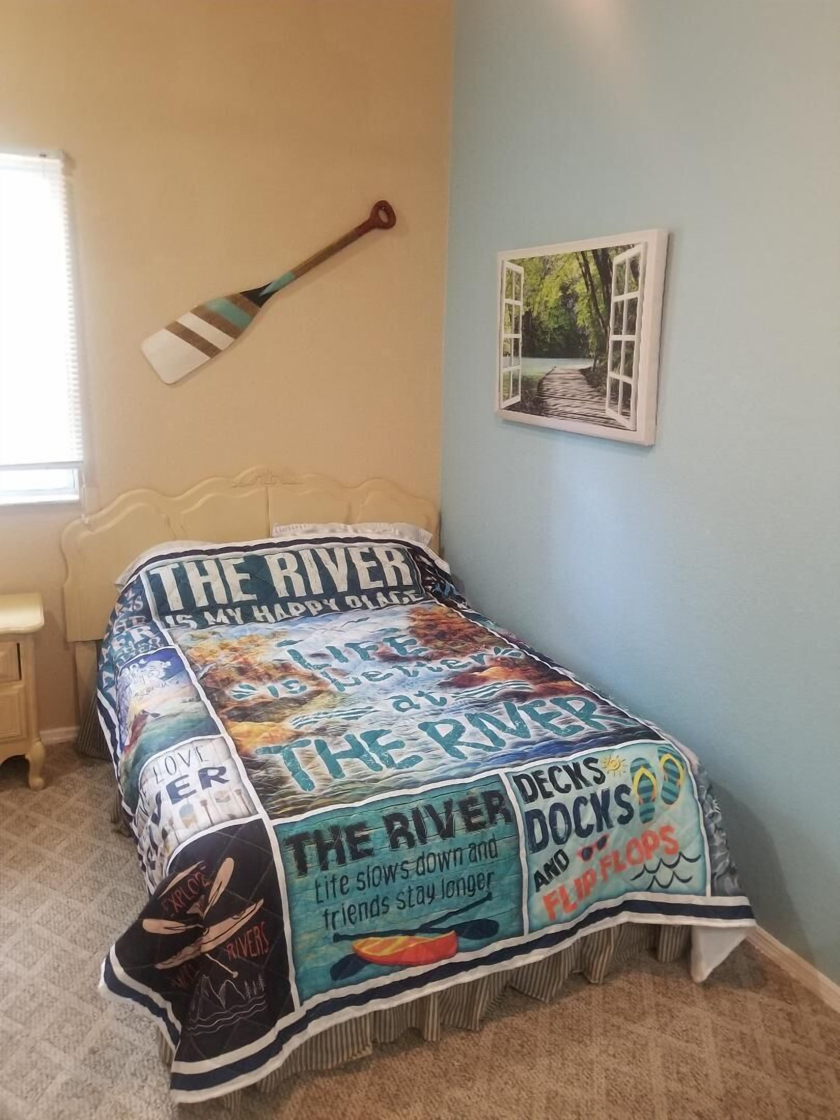 Ein Schlafzimmer mit einem Bett, das mit einer Decke mit Flussmuster bedeckt ist, einem Holzruder an der Wand und einem gerahmten Bild einer Flusslandschaft.