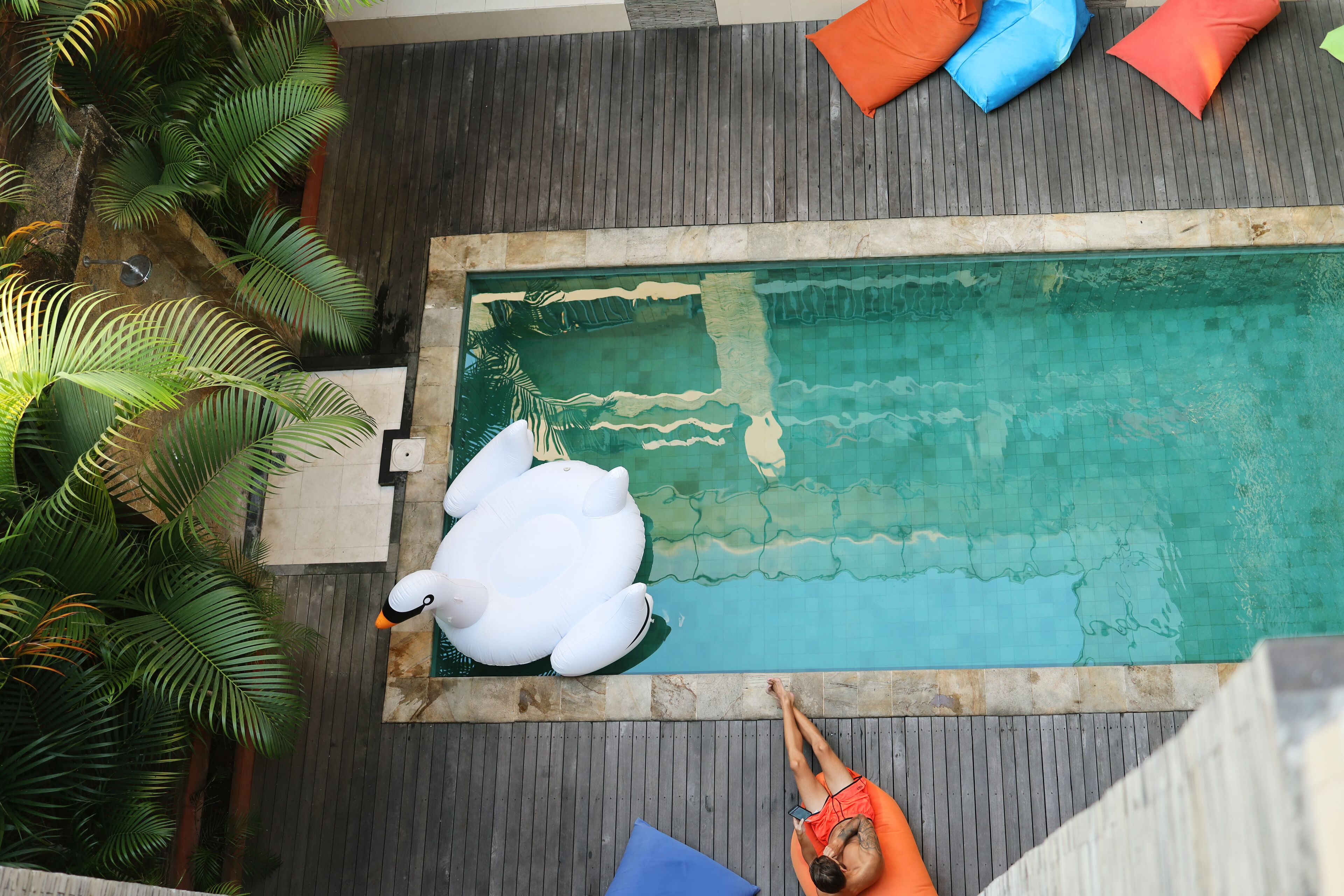 Une piscine sur le toit, avec un canard gonflable blanc, des palmiers et une personne se relaxant sur une chaise longue orange.