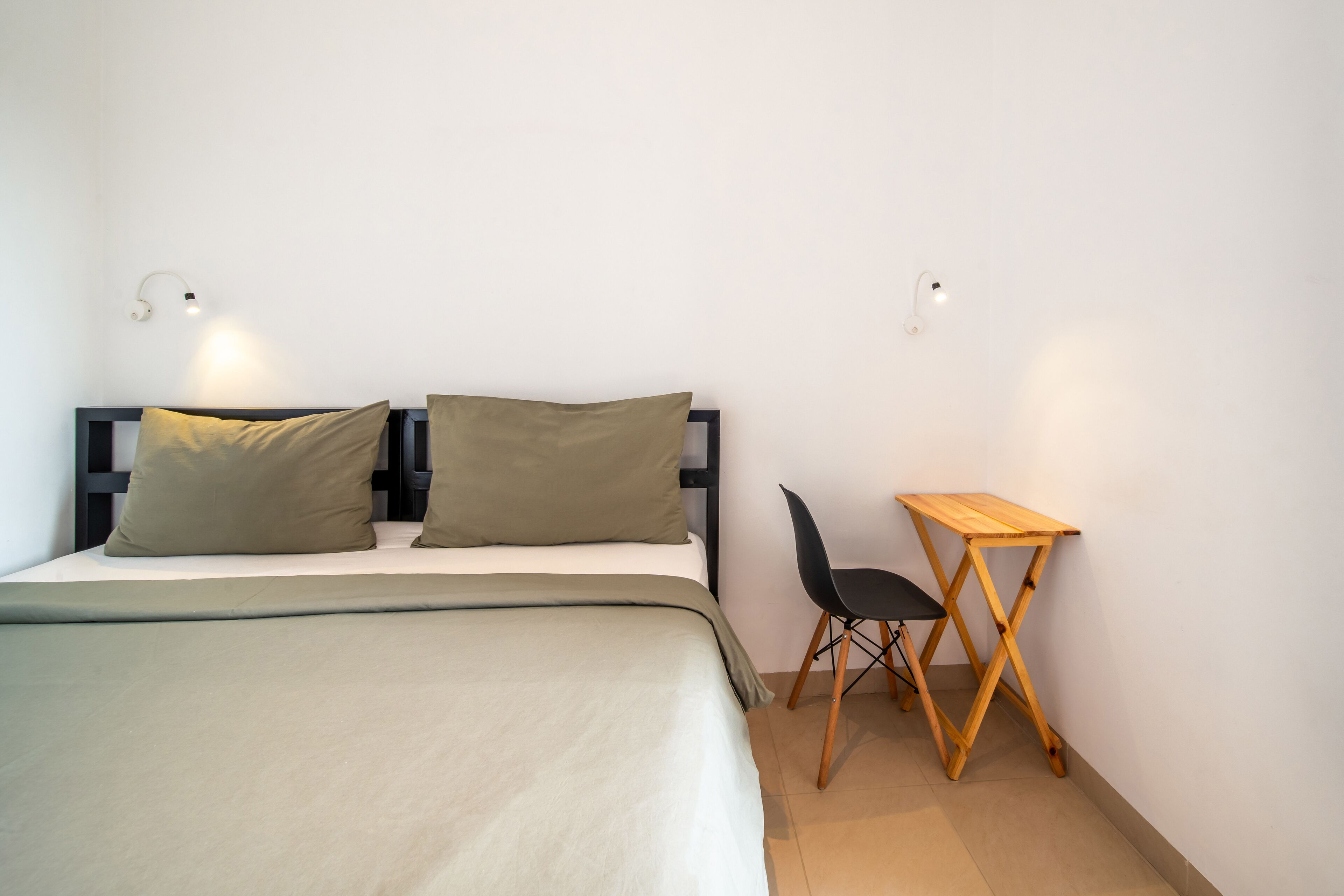 Un dormitorio con una cama, dos almohadas, un escritorio, una silla y dos luces fijadas a la pared.