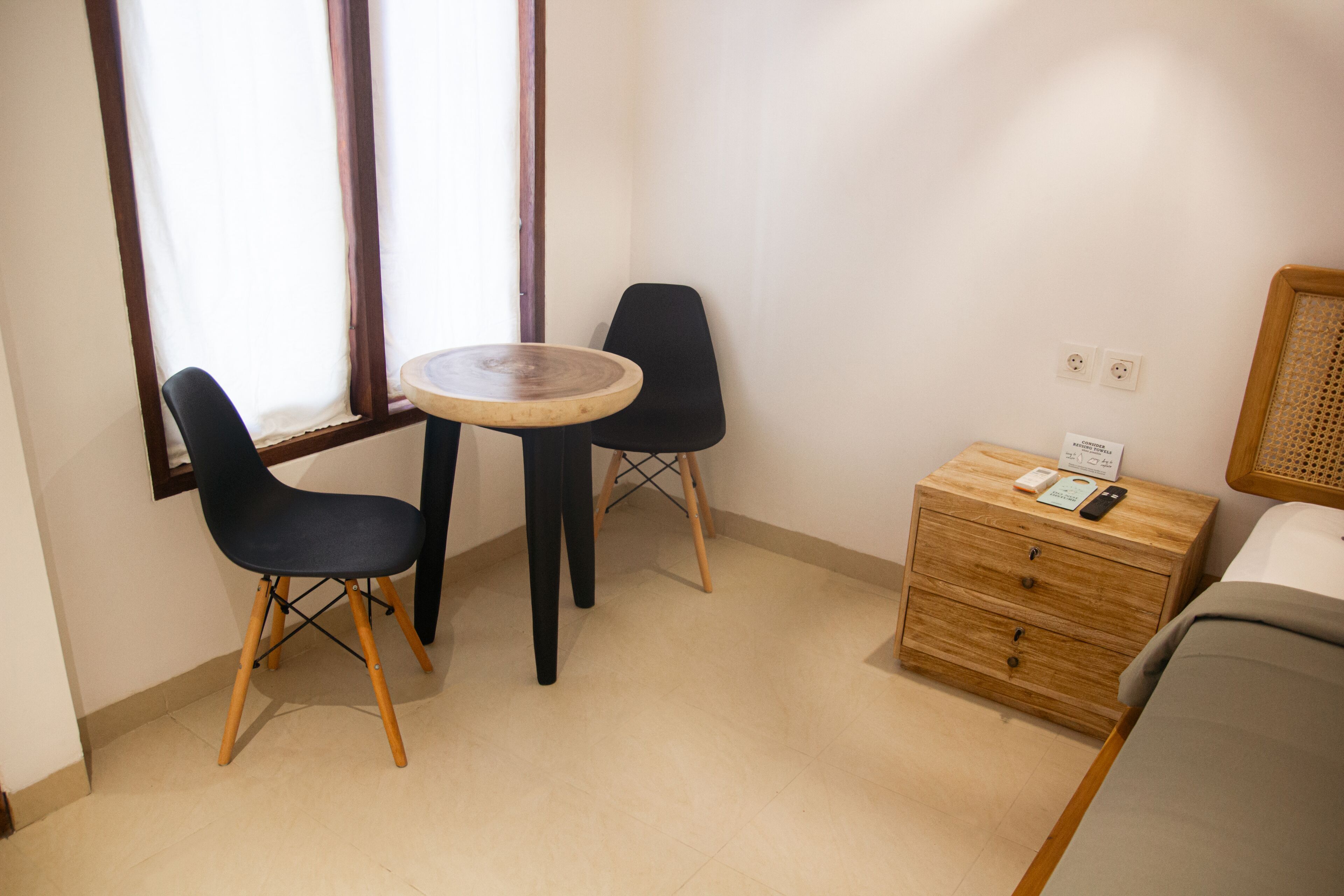 Ein kleiner, ordentlich eingerichteter Raum mit einem Bett, einem Nachttisch aus Holz, einem runden Tisch, zwei Stühlen und einem Fenster mit Vorhängen.