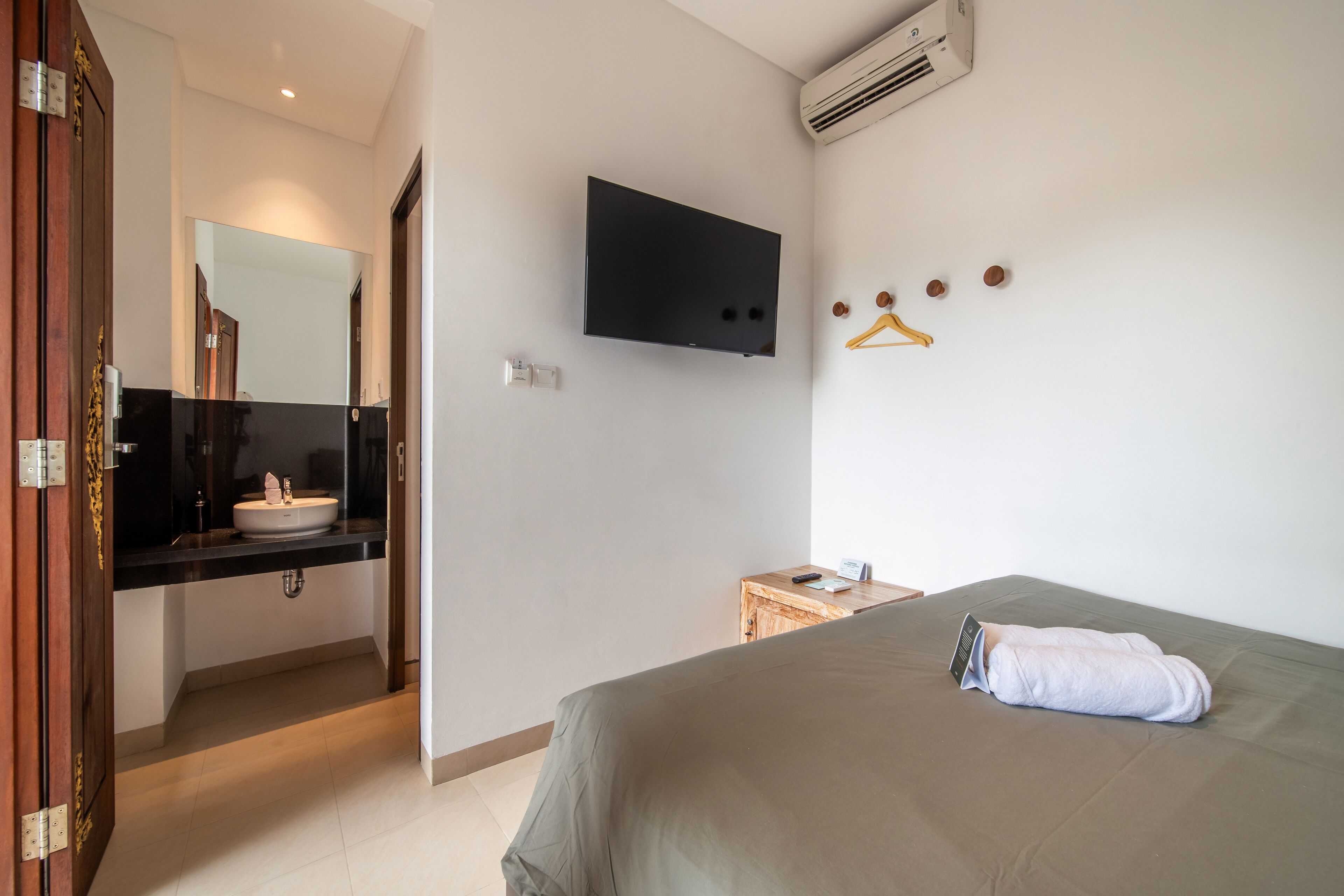 Habitació d'hotel amb un llit, televisió penjada a la paret, una petita taula de nit de fusta i un bany amb un lavabo i un mirall.