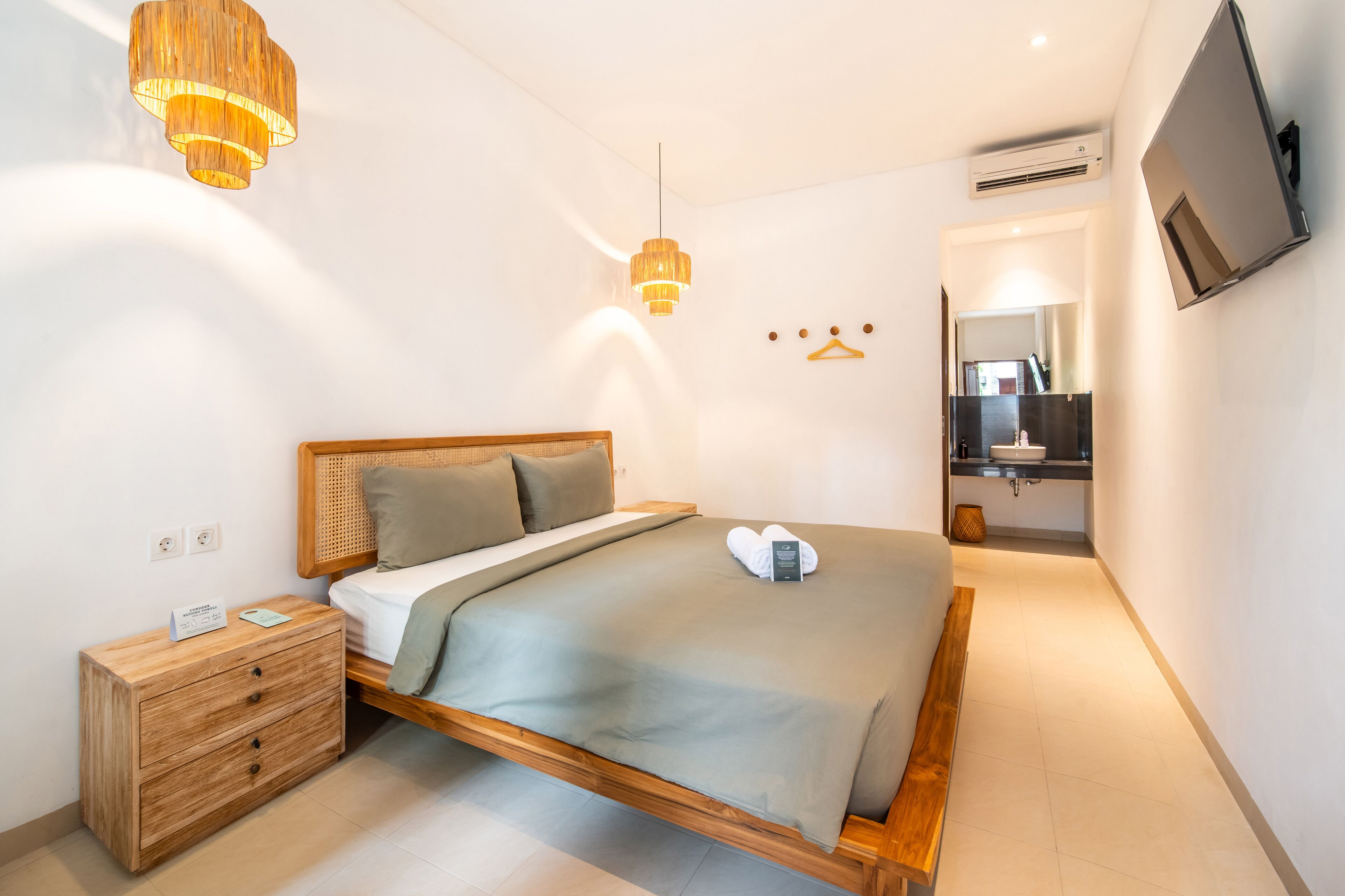 Un dormitorio moderno con cama de madera, mesitas de noche, lámpara colgante, televisor montado en la pared y baño visible a través de una puerta abierta.