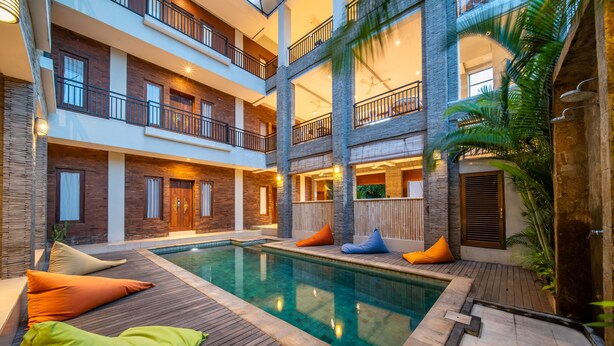 Uma área de piscina interior com vários níveis, deck de madeira, almofadas coloridas e piscina transparente.