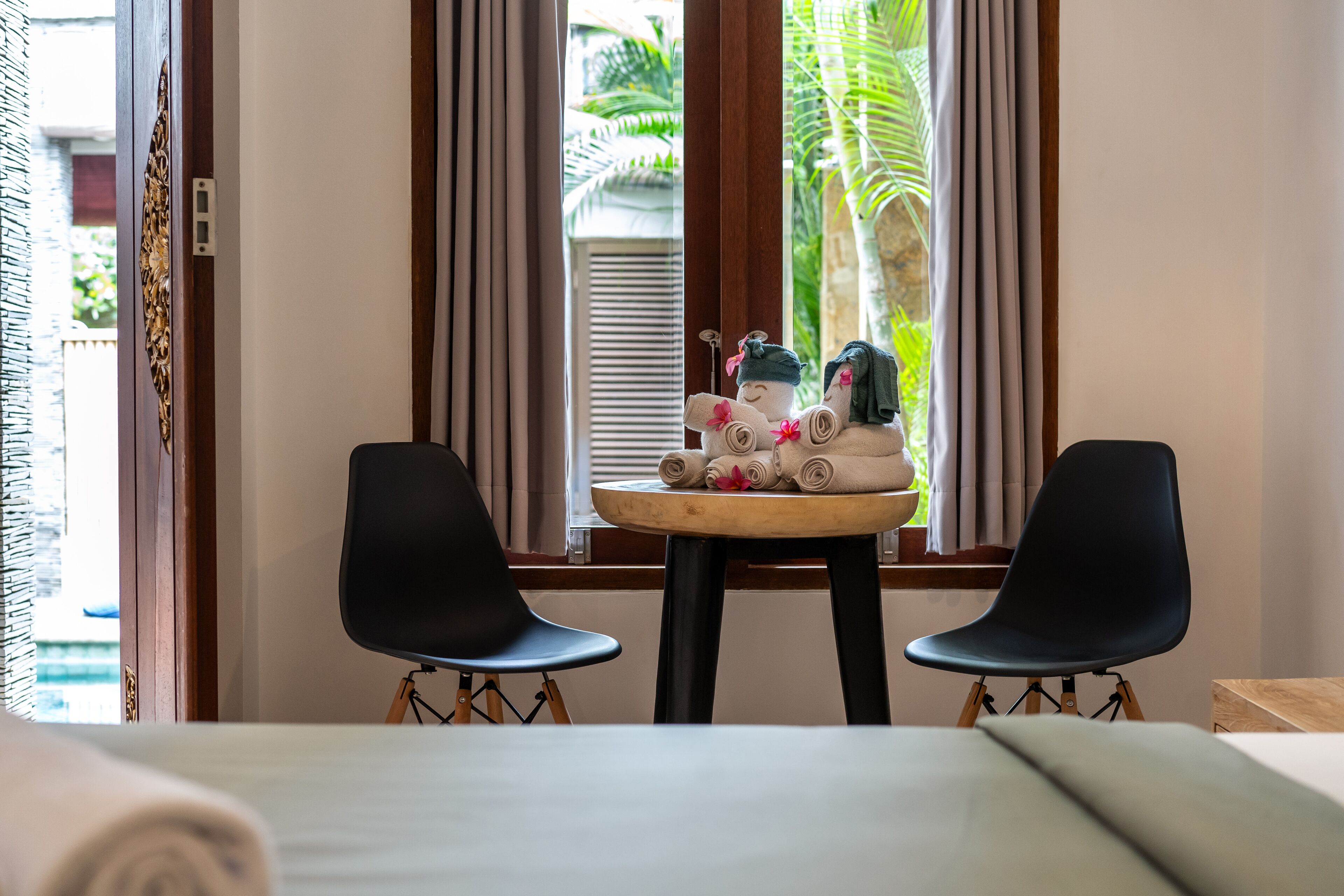 A room with two black chairs, a small round table with towels and toiletries, and a view of palm trees through a window with curtains.