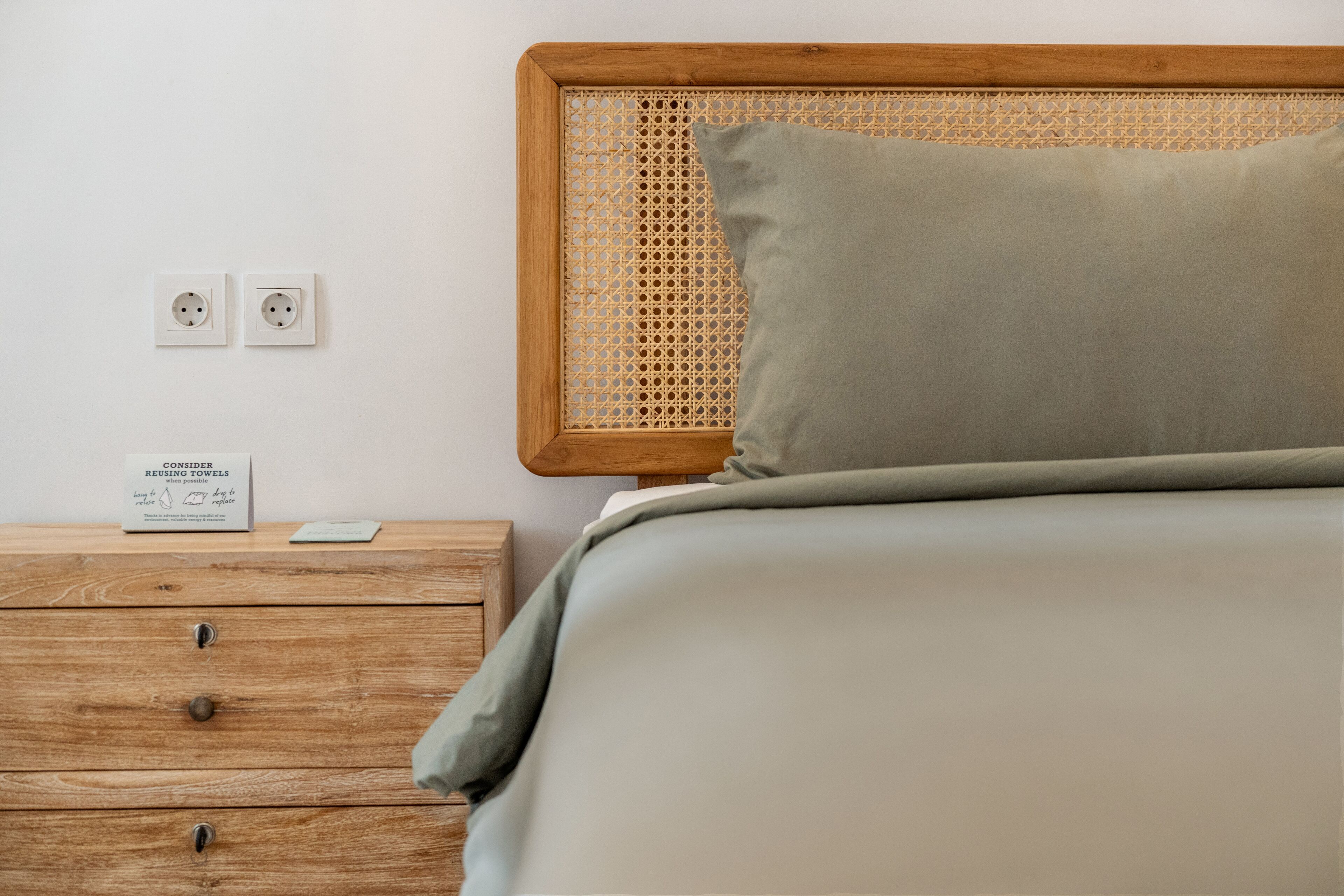A neatly made bed with a wooden headboard and a single pillow, a wooden nightstand with drawers, and two electrical outlets on the wall.