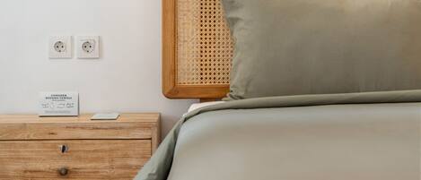 A neatly made bed with a wooden headboard and a single pillow, a wooden nightstand with drawers, and two electrical outlets on the wall.