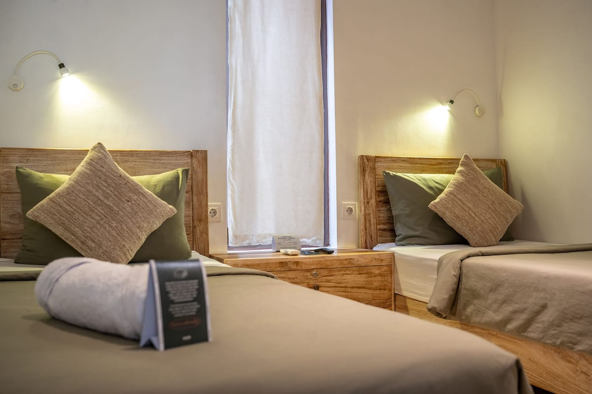 A hotel room with two beds, each with a wooden headboard, a nightstand, and a bedside table. There is a window with a curtain, and the room has a neutral color scheme.