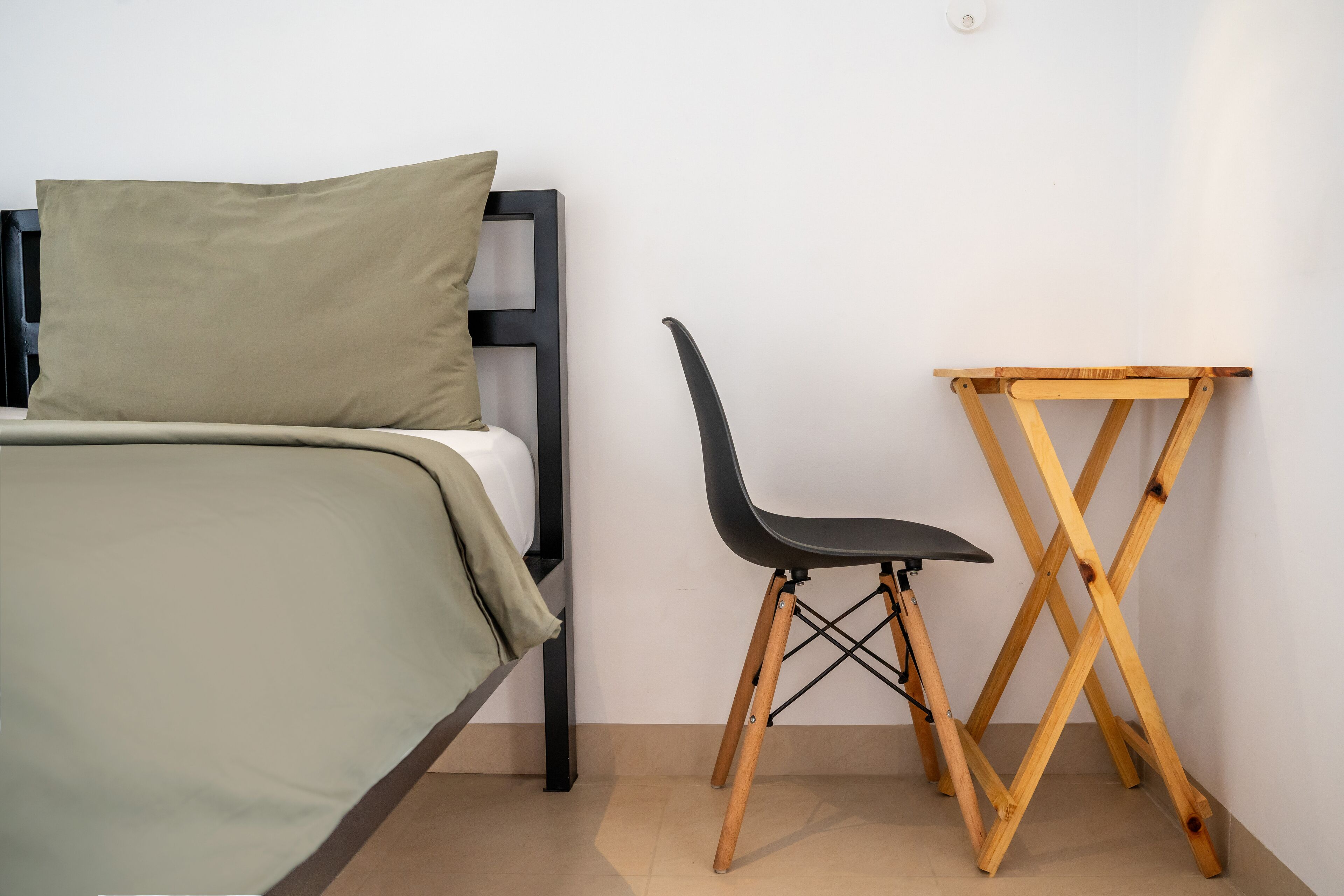 Una cama con colcha verde y cabecera negra, una silla negra y una mesa de madera frente a una pared lisa.