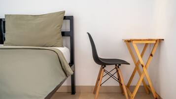 Una cama con colcha verde y cabecera negra, una silla negra y una mesa de madera frente a una pared lisa.