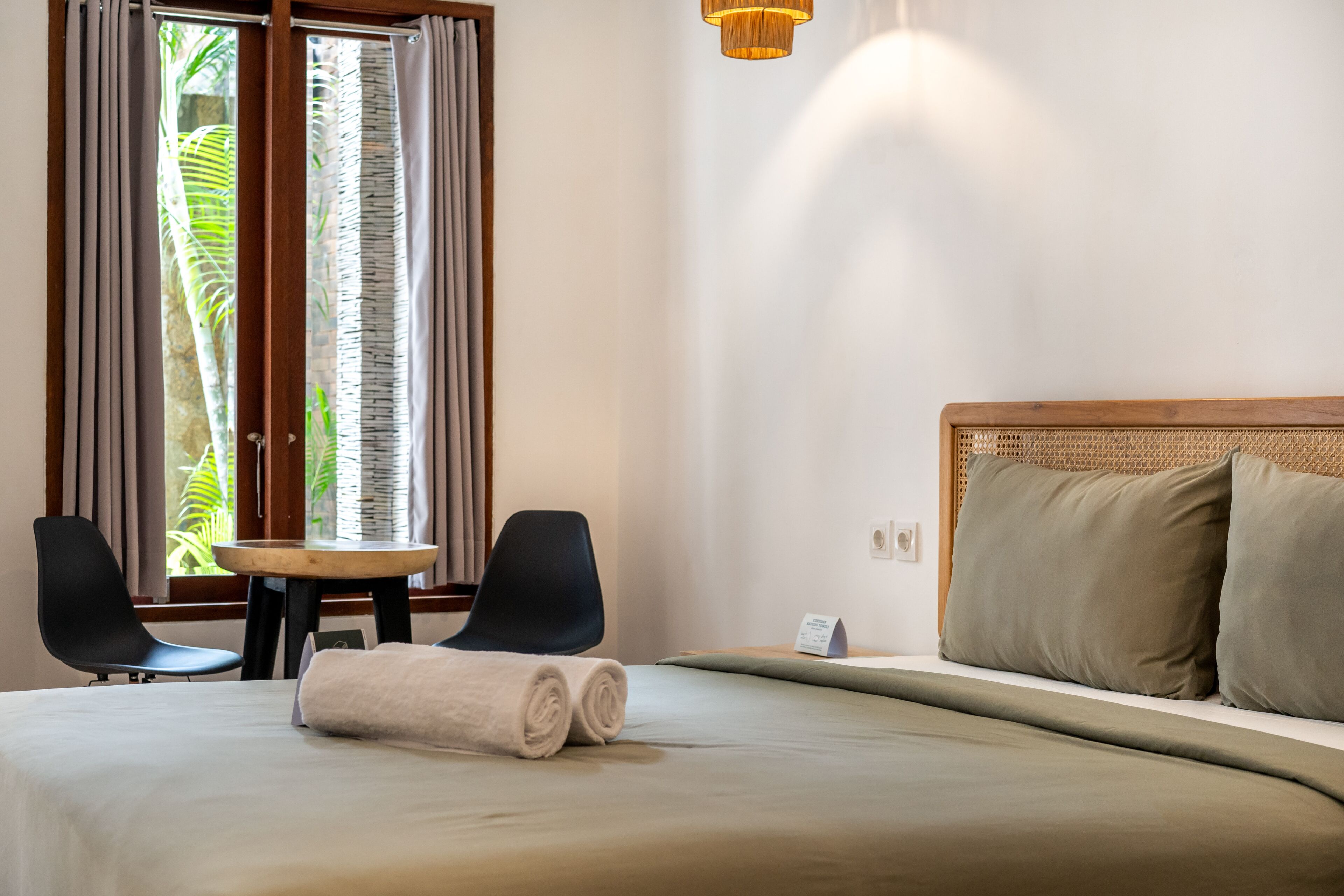 A hotel room with a bed, two chairs, a small table, and a view of greenery through the window.