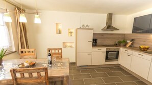 A modern kitchen with white cabinets, a built-in oven, and a coffee maker. There is a dining table with chairs and a potted plant. - Wildnisresidenz am Nationalpark (Lindberg)
