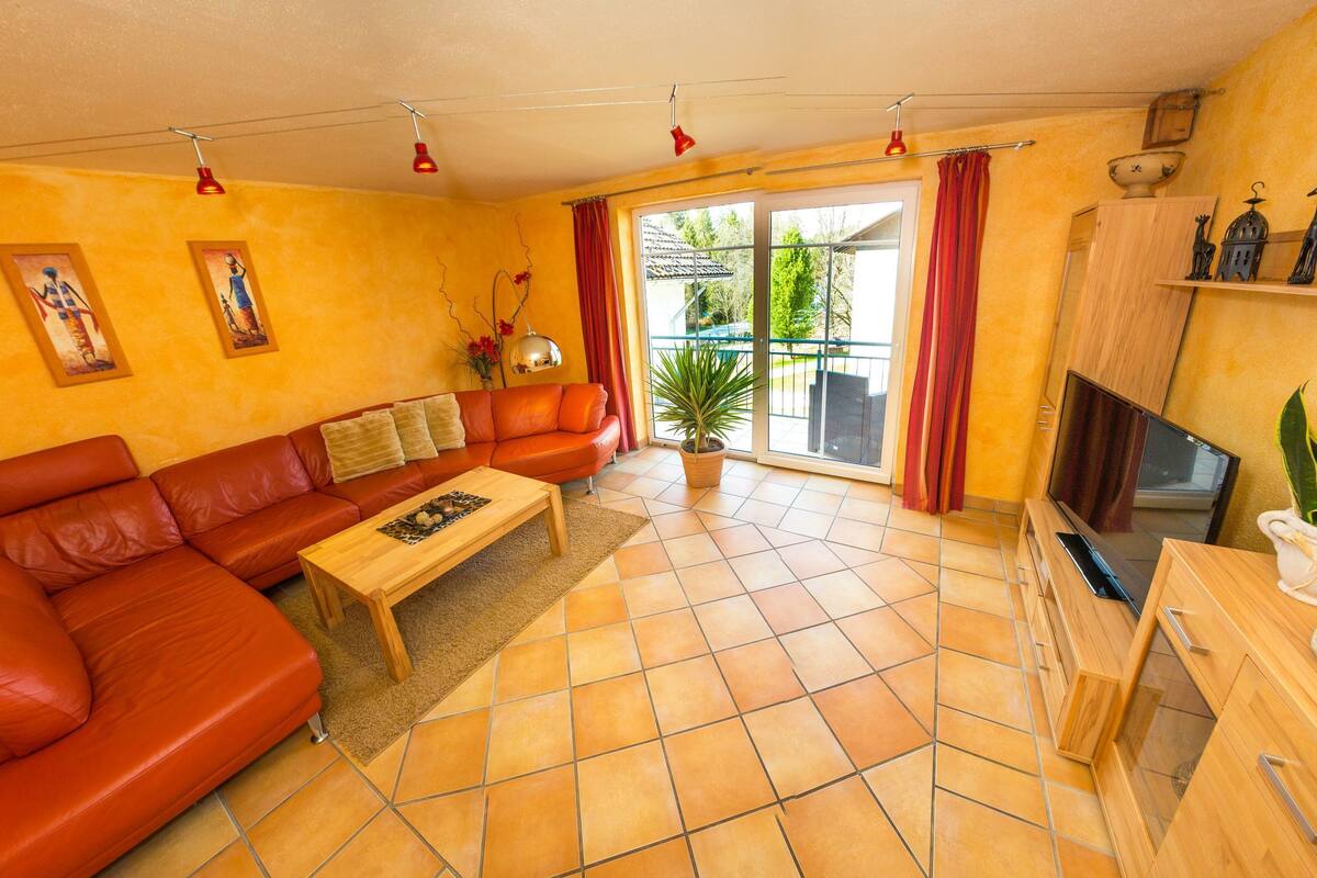Un salón con un sofá de cuero rojo, una mesa de centro de madera y un televisor sobre un mueble de madera. Hay una planta en maceta y un cuadro en la pared.