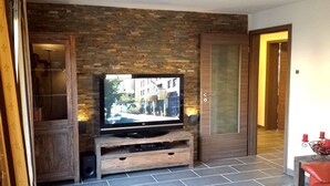 A living room with a television on a wooden stand, a brick wall, and a door leading to another room. - Wildnisresidenz am Nationalpark (Lindberg)