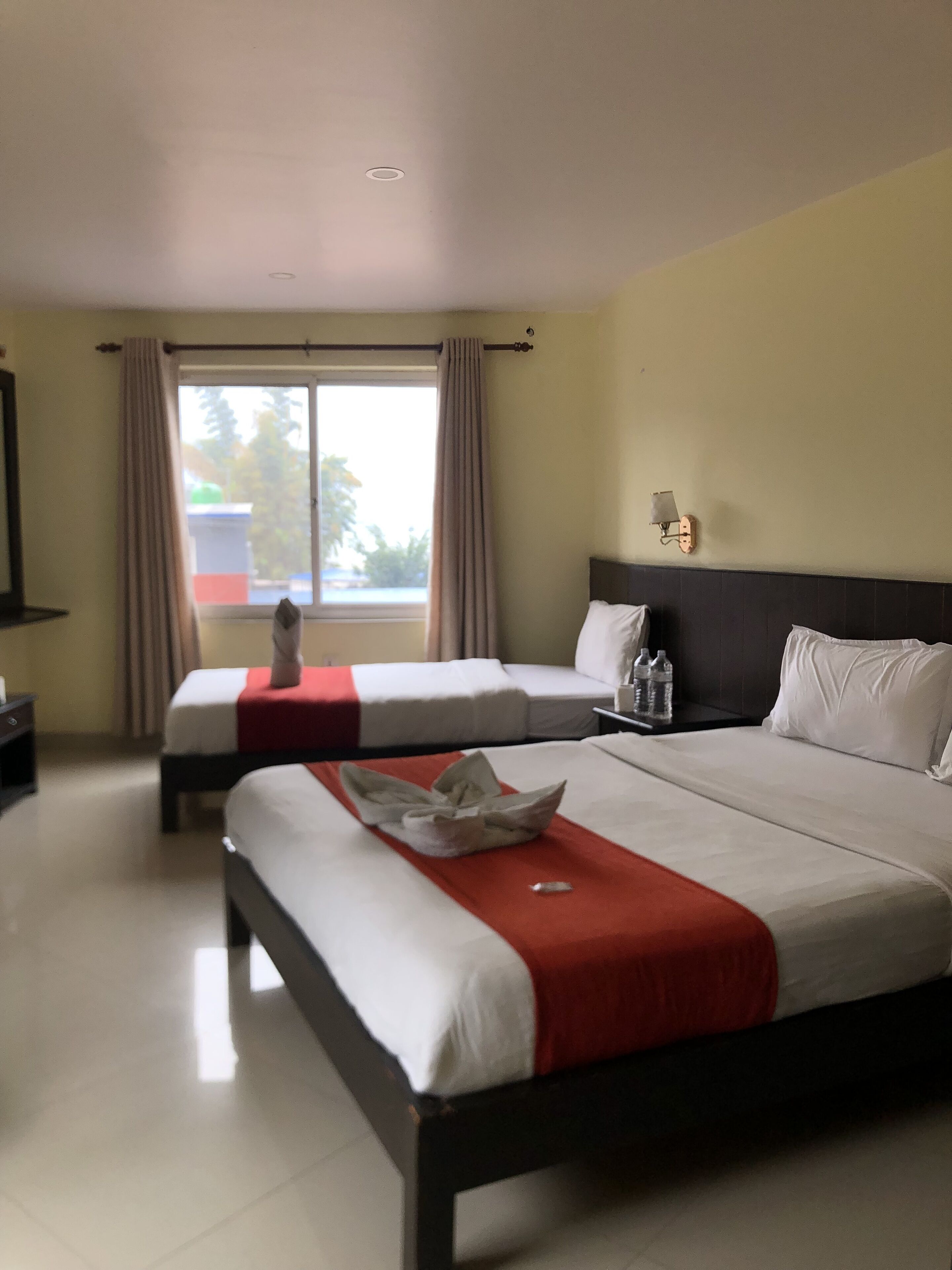 Habitación de hotel con dos camas, una ventana con cortinas, una mesita de noche con lámpara y vistas al exterior.