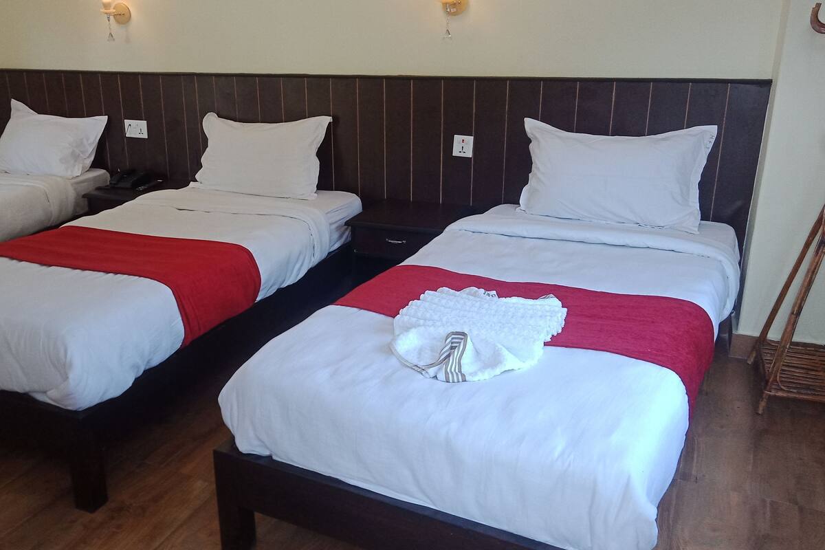 A hotel room with three single beds, each with white bedding and red accents, wooden headboards, and bedside tables.