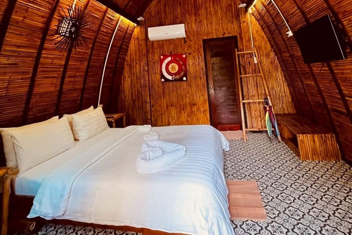 A cozy, rustic room with a high, curved wooden ceiling, a bed with neatly arranged pillows, a small ladder, and a mounted TV.