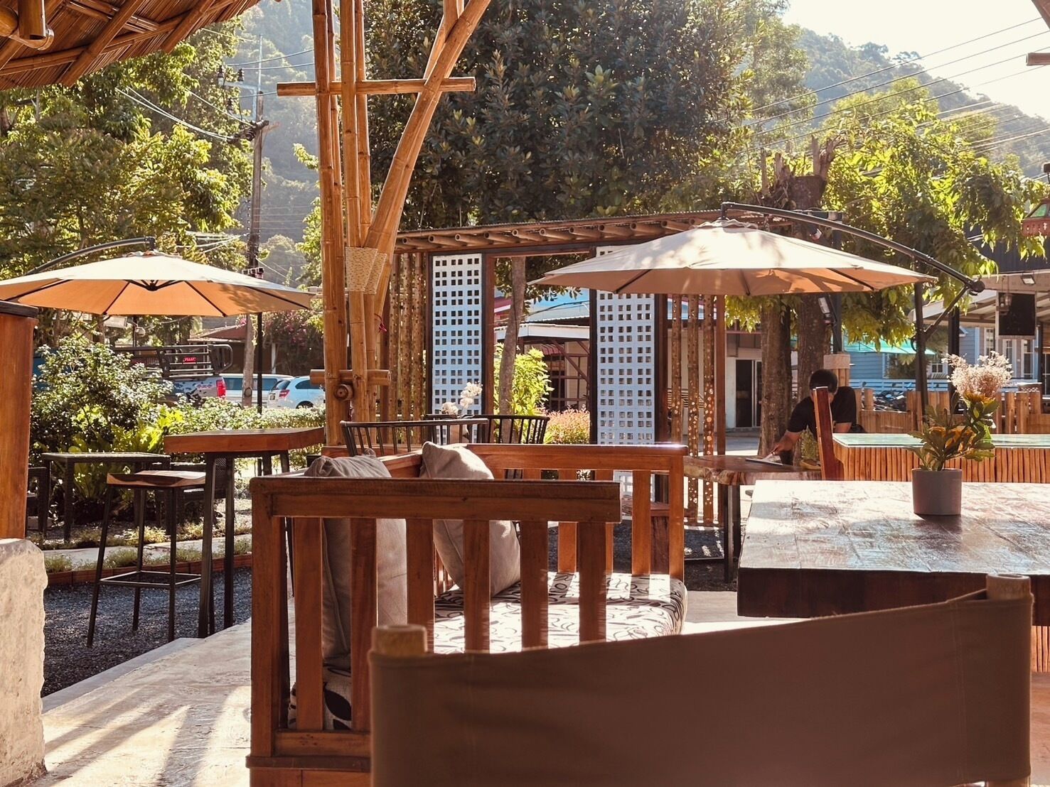 An outdoor seating area with wooden furniture and umbrellas, surrounded by greenery and a bamboo structure.