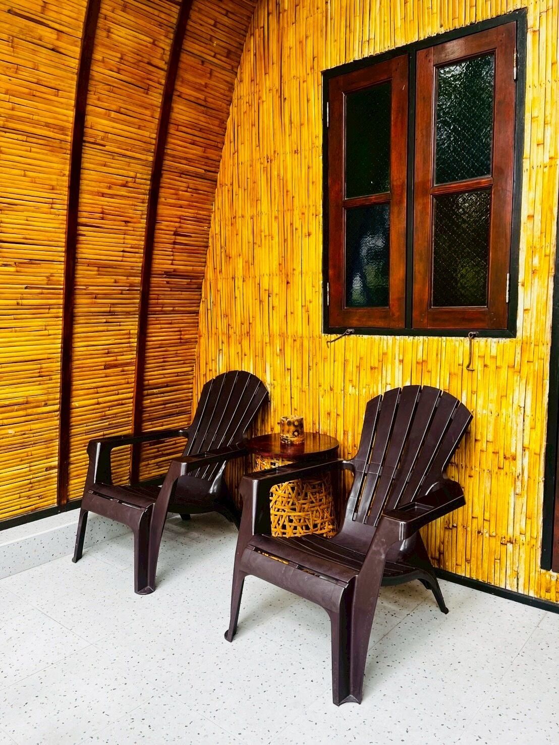 Una habitación con una pared de madera curvada y una ventana con marcos de madera oscura. Hay dos sillas de madera oscura y una mesita redonda con una cesta tejida.
