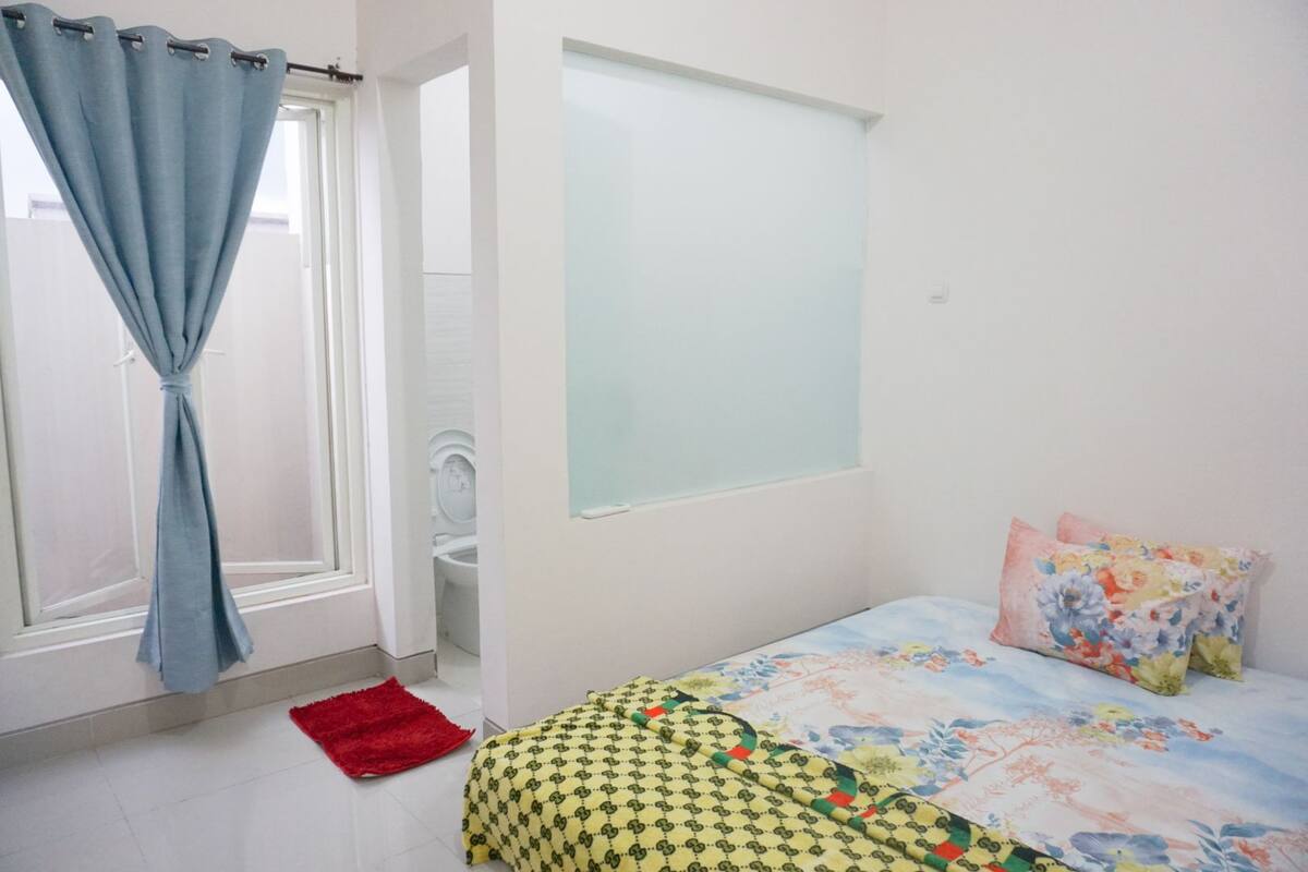 A bedroom with a bed, a window with curtains, a toilet, and a red rug.