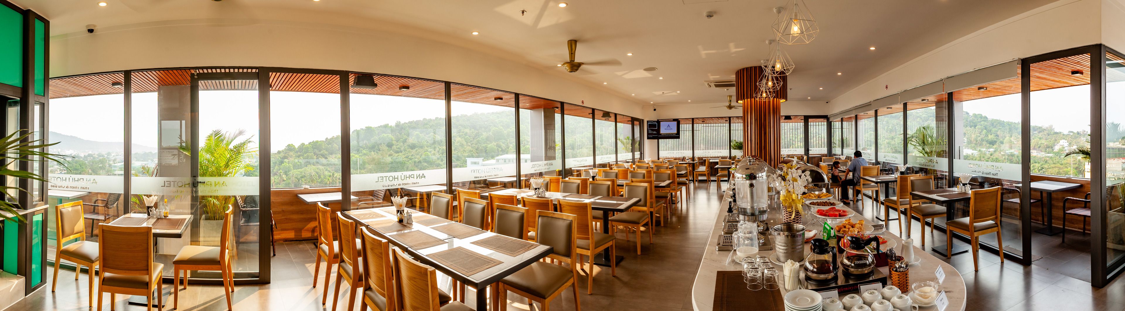 A spacious restaurant with high ceilings and large windows offering views of the surrounding landscape. Tables are neatly arranged with chairs, and a buffet line is visible in the center.