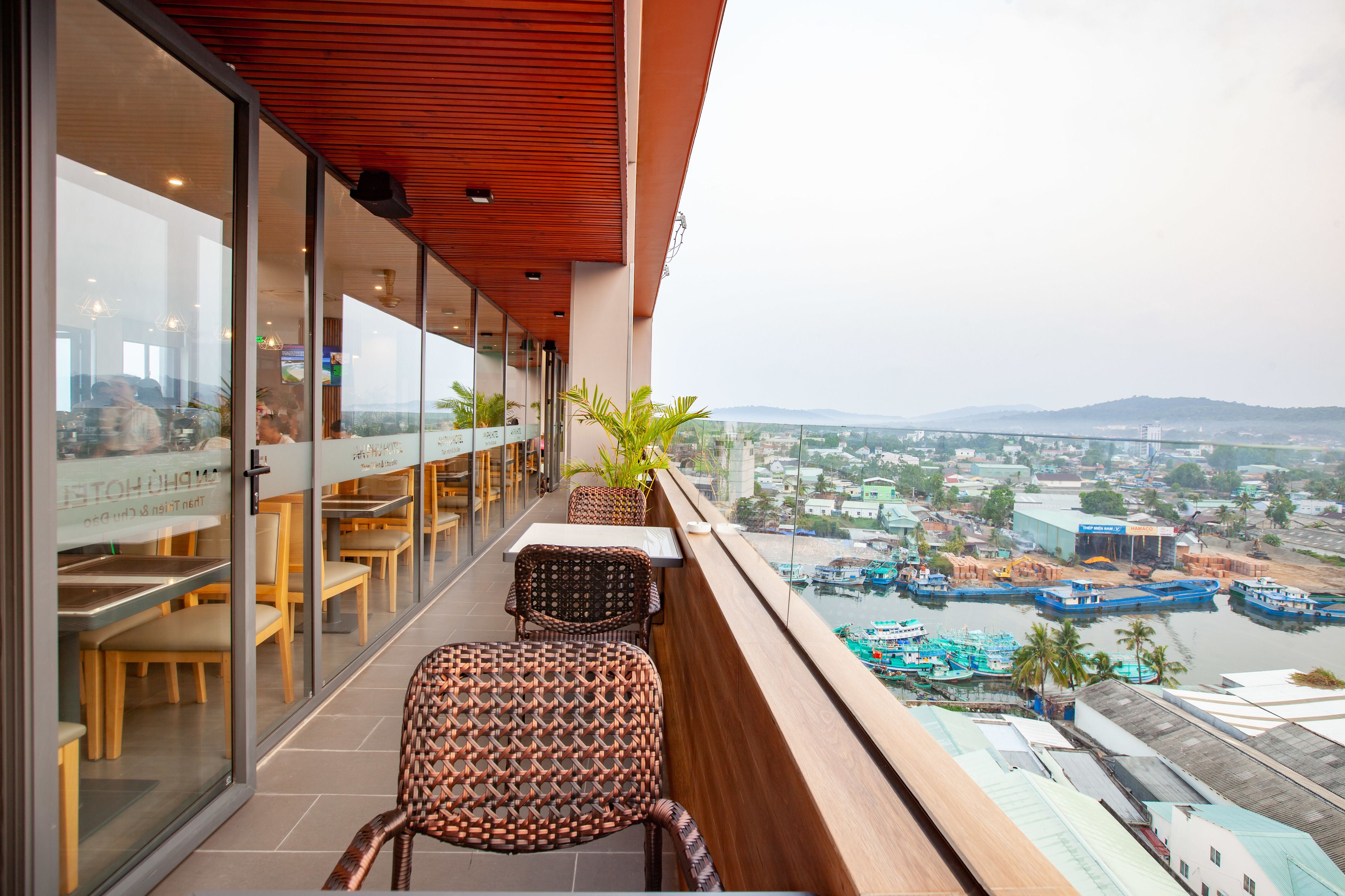 A balcony with wicker chairs and a table, overlooking a river with boats and industrial buildings.