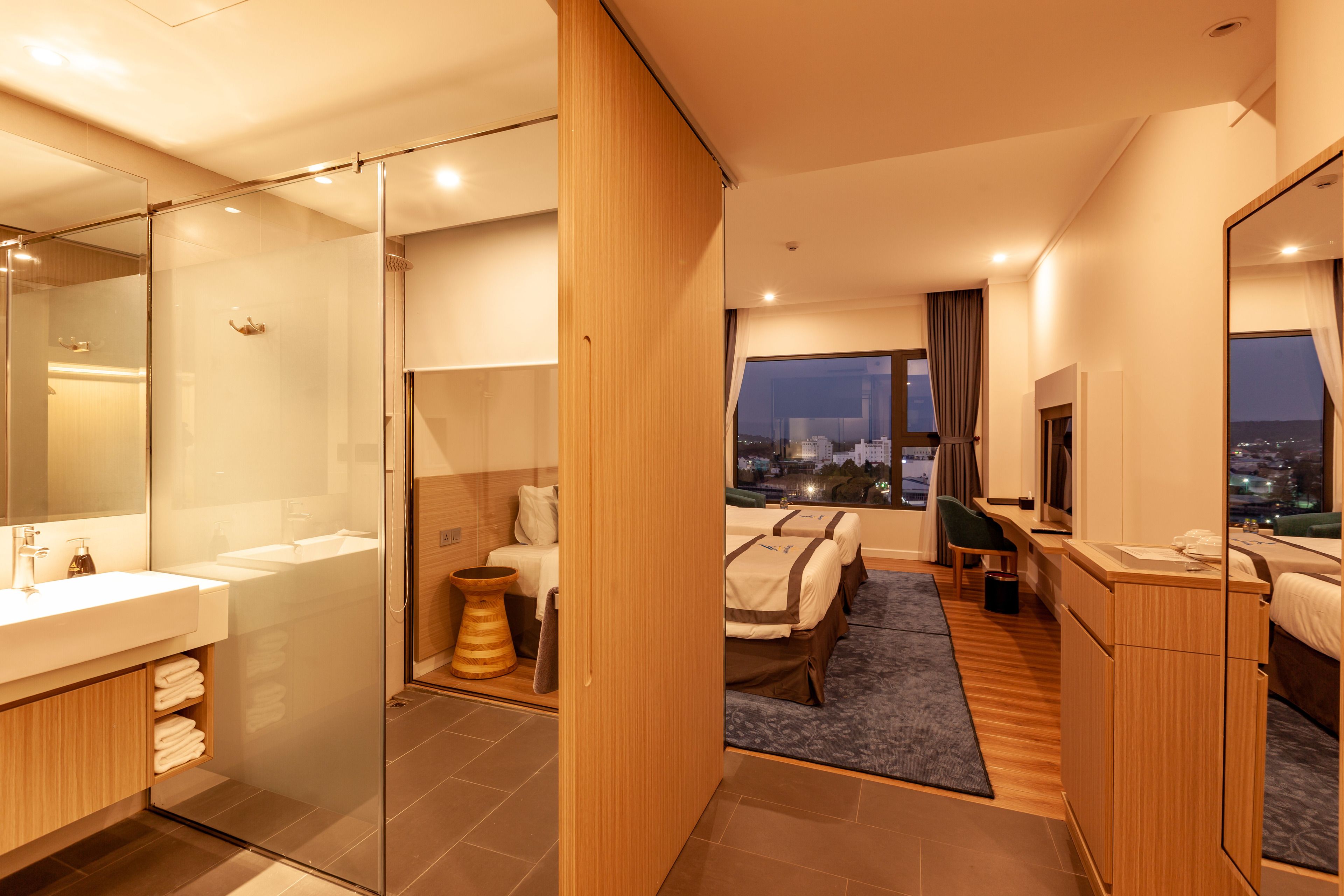 Una habitación de hotel moderna con cabina de ducha de vidrio, una cama grande con vistas a la ciudad de noche, una zona de estar separada y un tocador de madera.