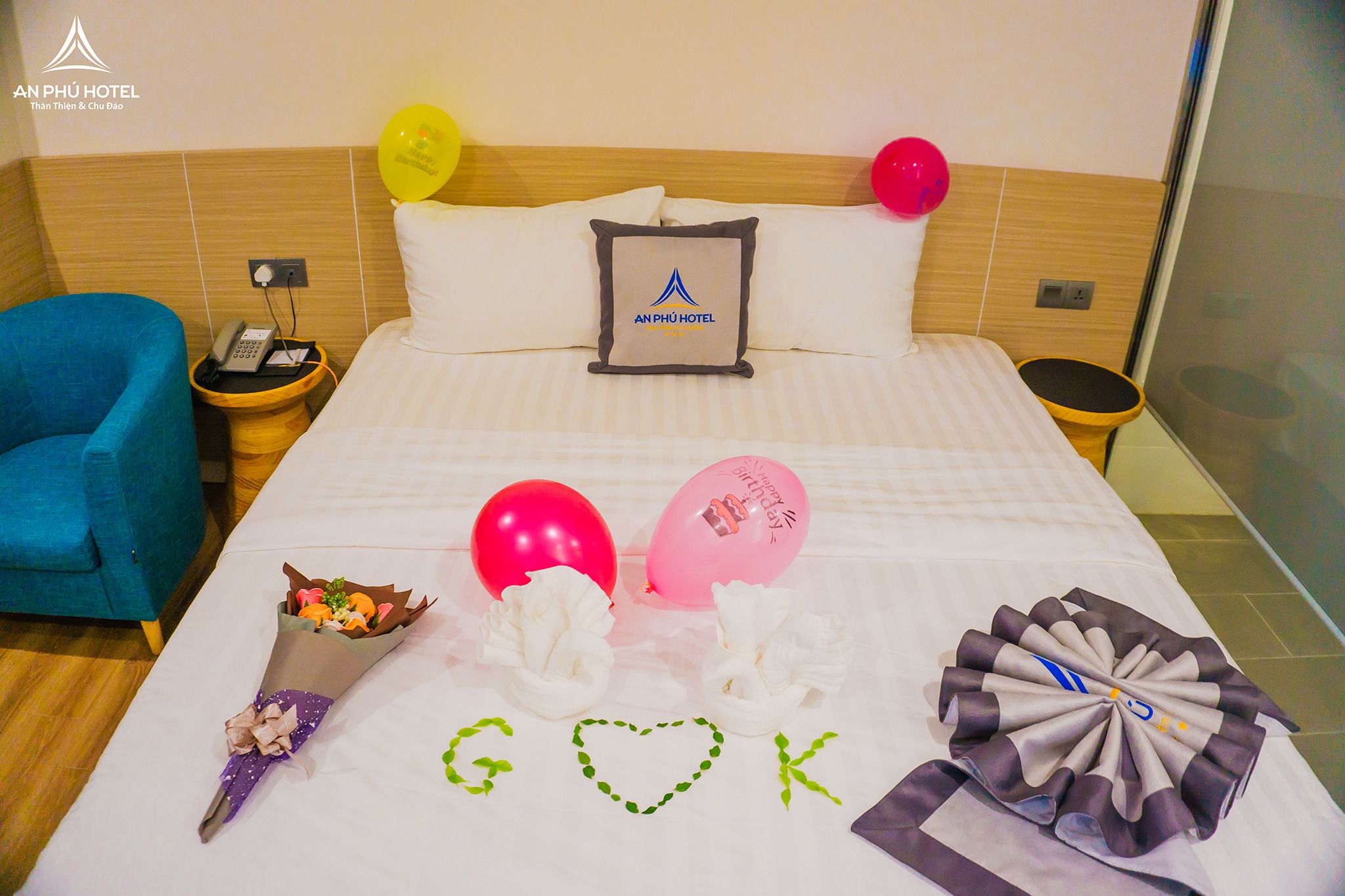 A hotel room with a bed, a nightstand, a blue chair, balloons, and a bouquet of flowers.