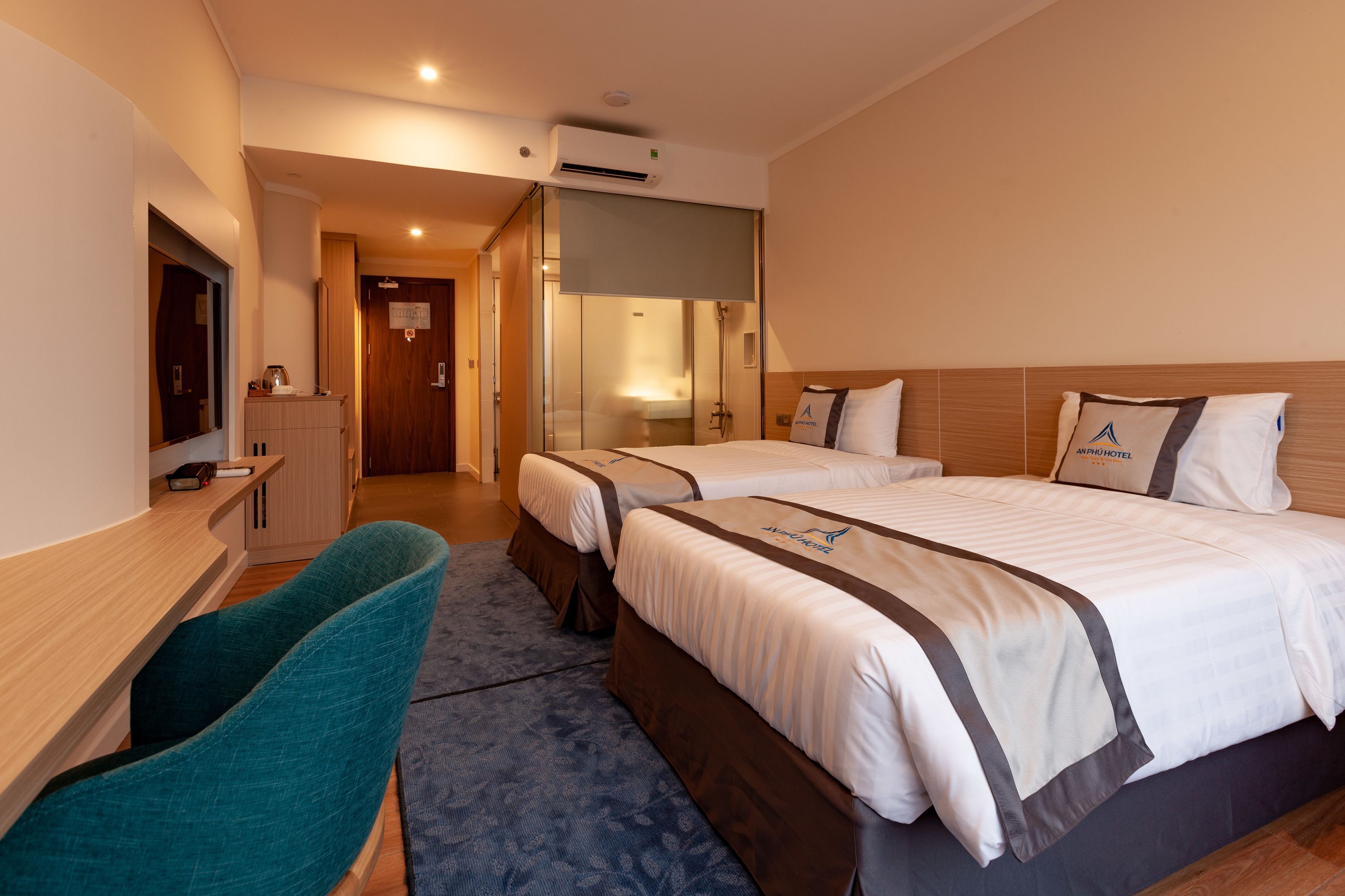 A hotel room with two beds, a desk, a chair, and a bathroom visible through a glass door.