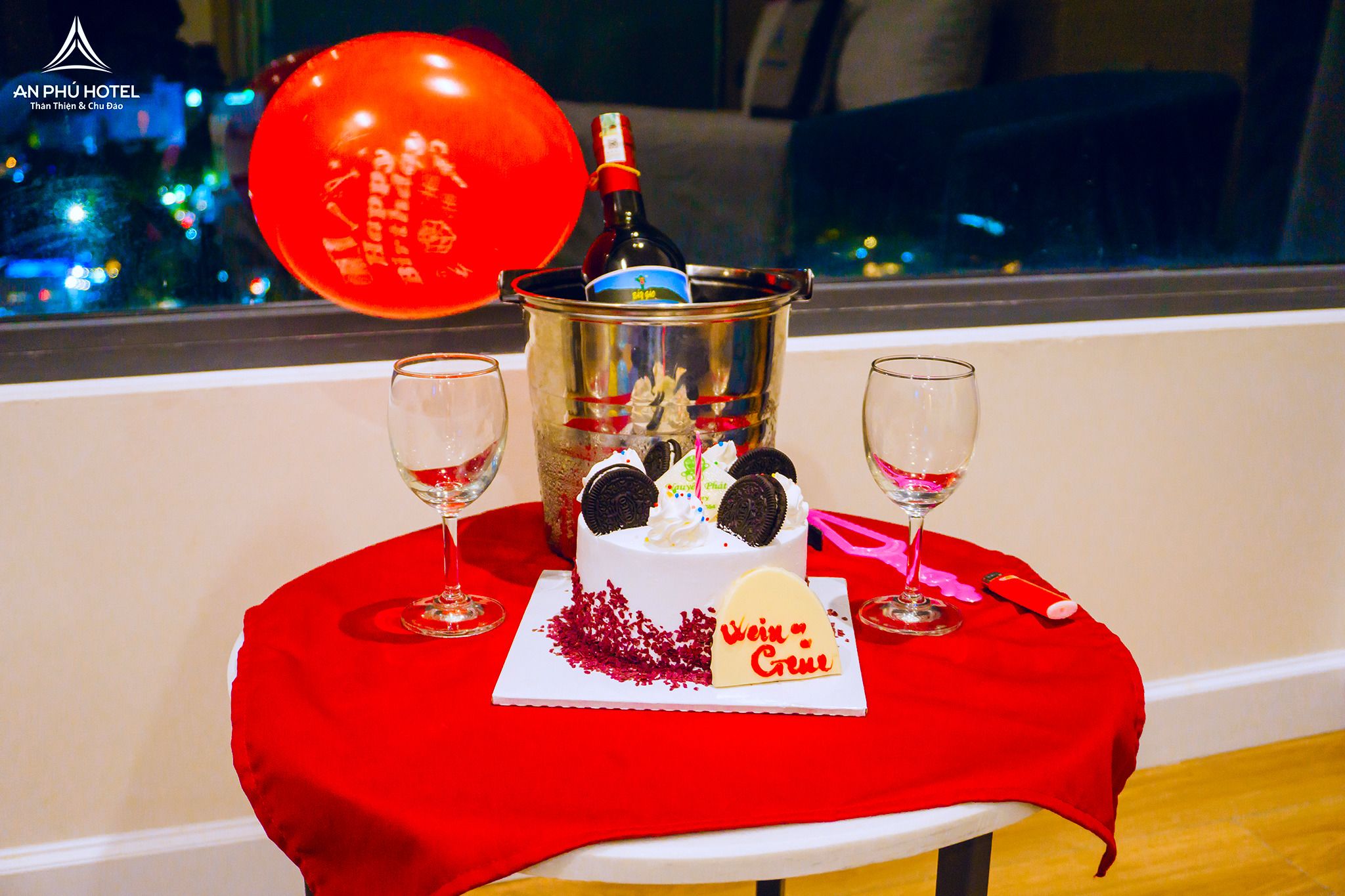 A table with a birthday cake, a bottle of champagne, and two glasses on a red tablecloth.