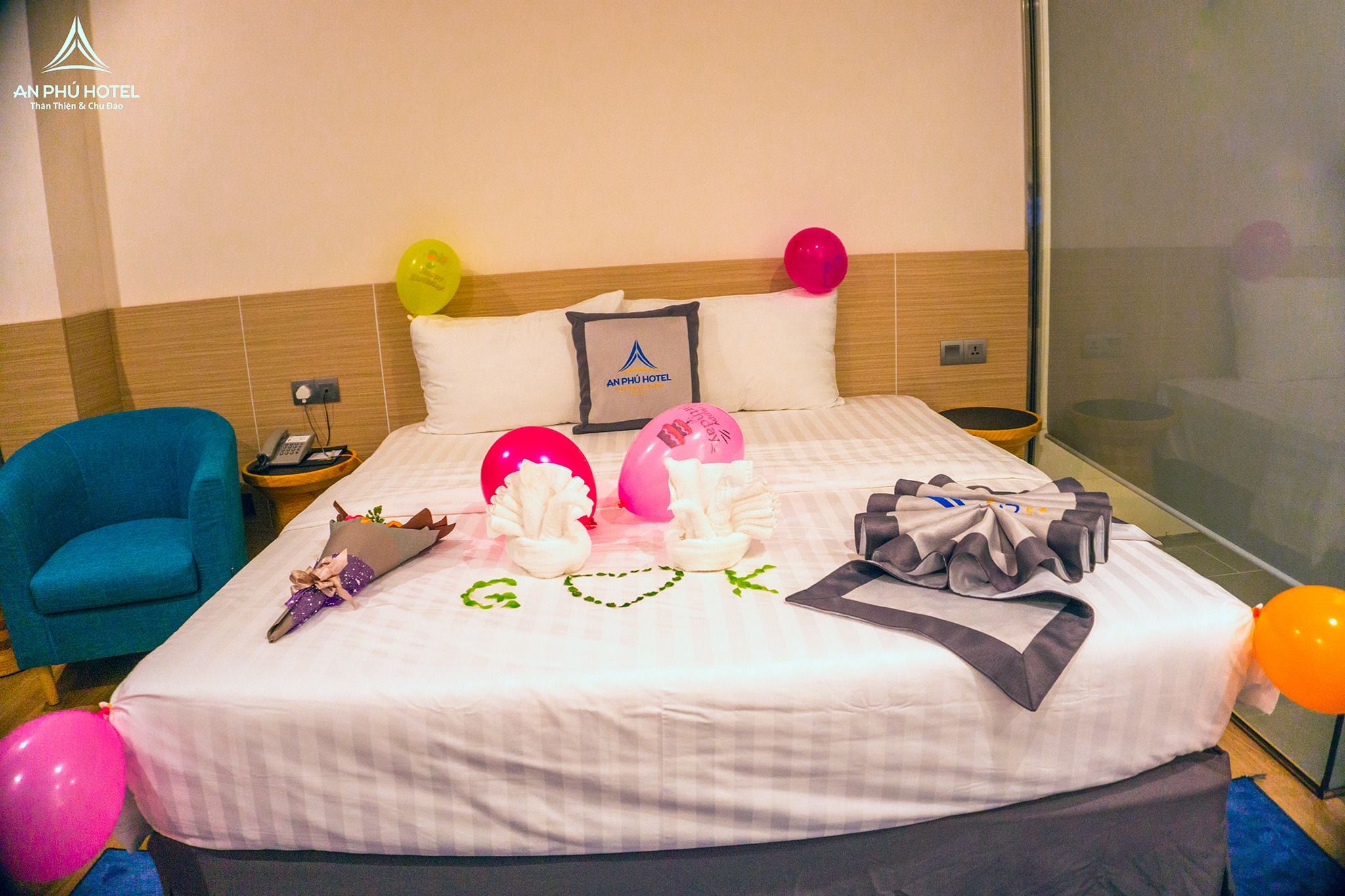 Una habitación de hotel con cama, un sillón azul, una mesita de noche con teléfono y globos.