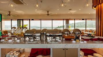 A buffet setup with multiple food stations, a ceiling fan, and large windows offering a scenic view.