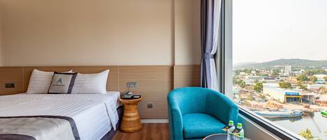 Ein Hotelzimmer mit einem Bett, einem blauen Sessel, einem kleinen Tisch mit Flaschen und Blick auf eine Stadtlandschaft durch ein großes Fenster.