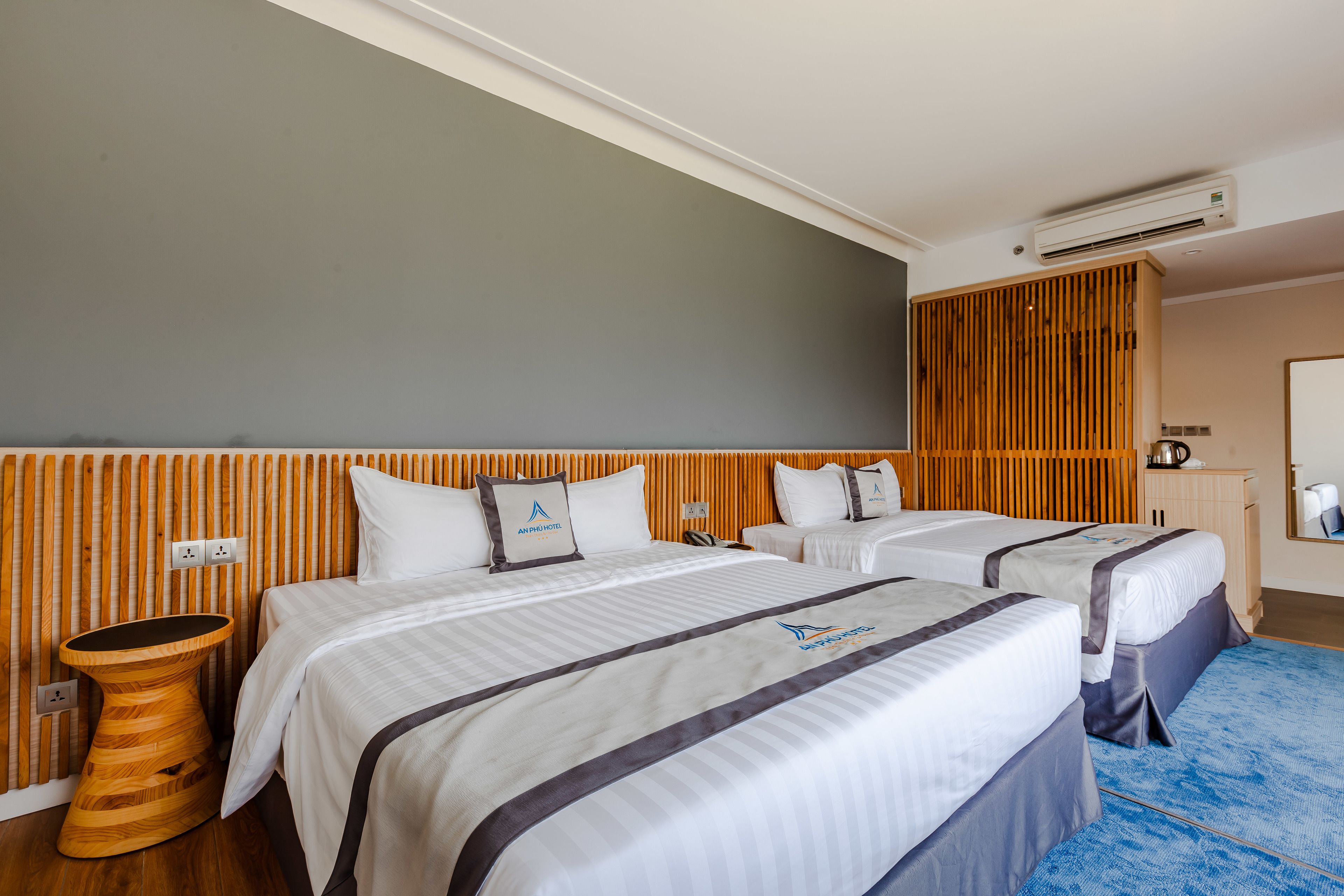 A hotel room with two beds, a wooden headboard, a round wooden side table, and a blue carpet.