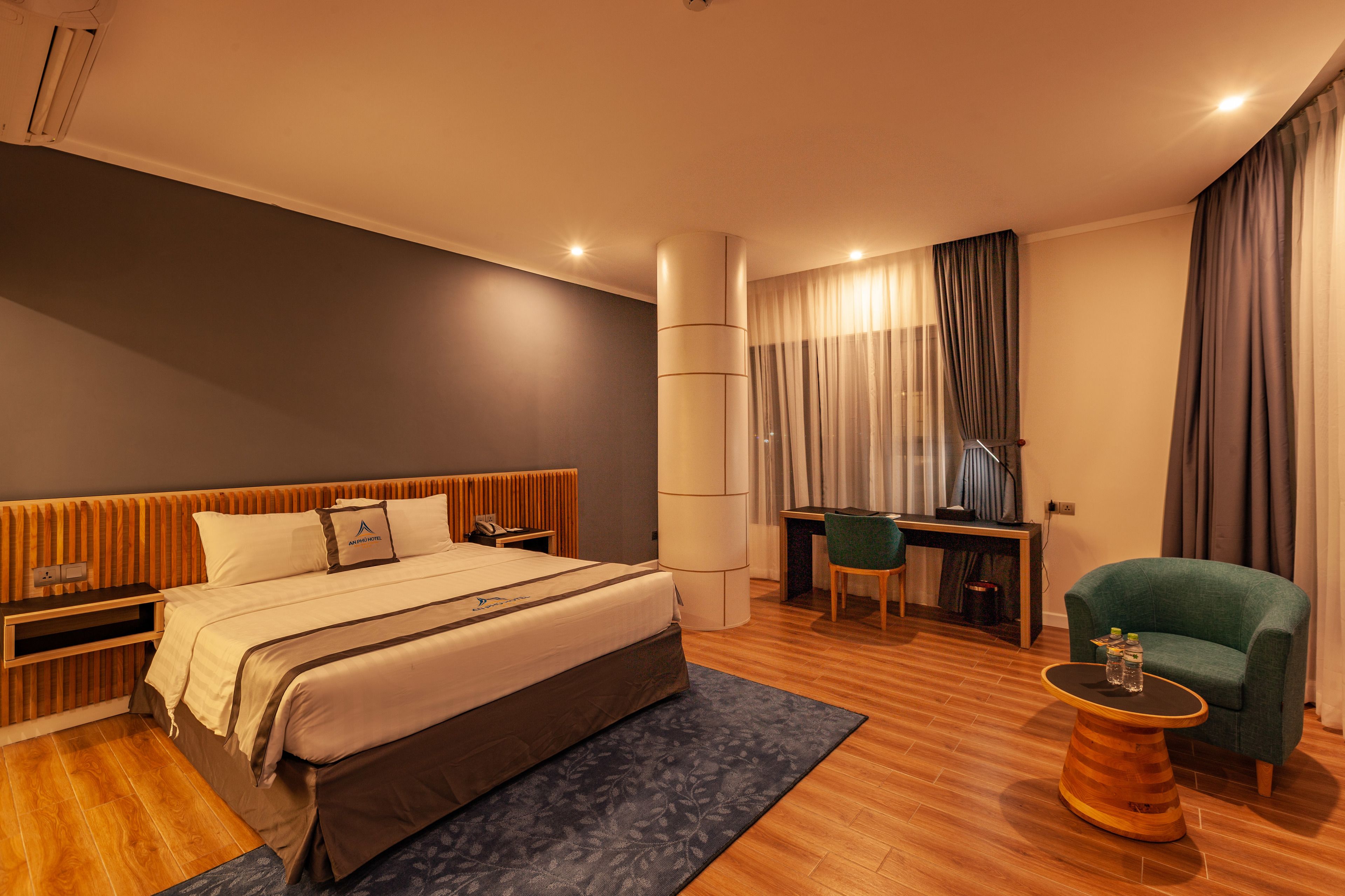 A modern hotel room with a large bed, a desk, a chair, and a small round table with a bottle.