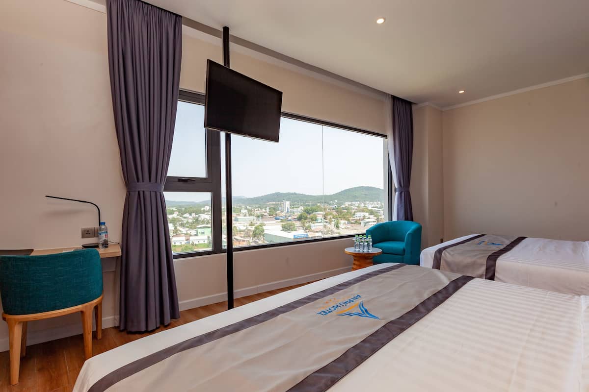 Habitación de hotel con dos camas, un escritorio con silla, televisor montado en la pared y vistas a un paisaje urbano a través de amplias ventanas.