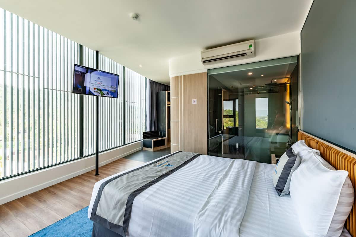 Una habitación de hotel moderna con una cama grande, un televisor montado en la pared y un ventanal corredero que da a un balcón.