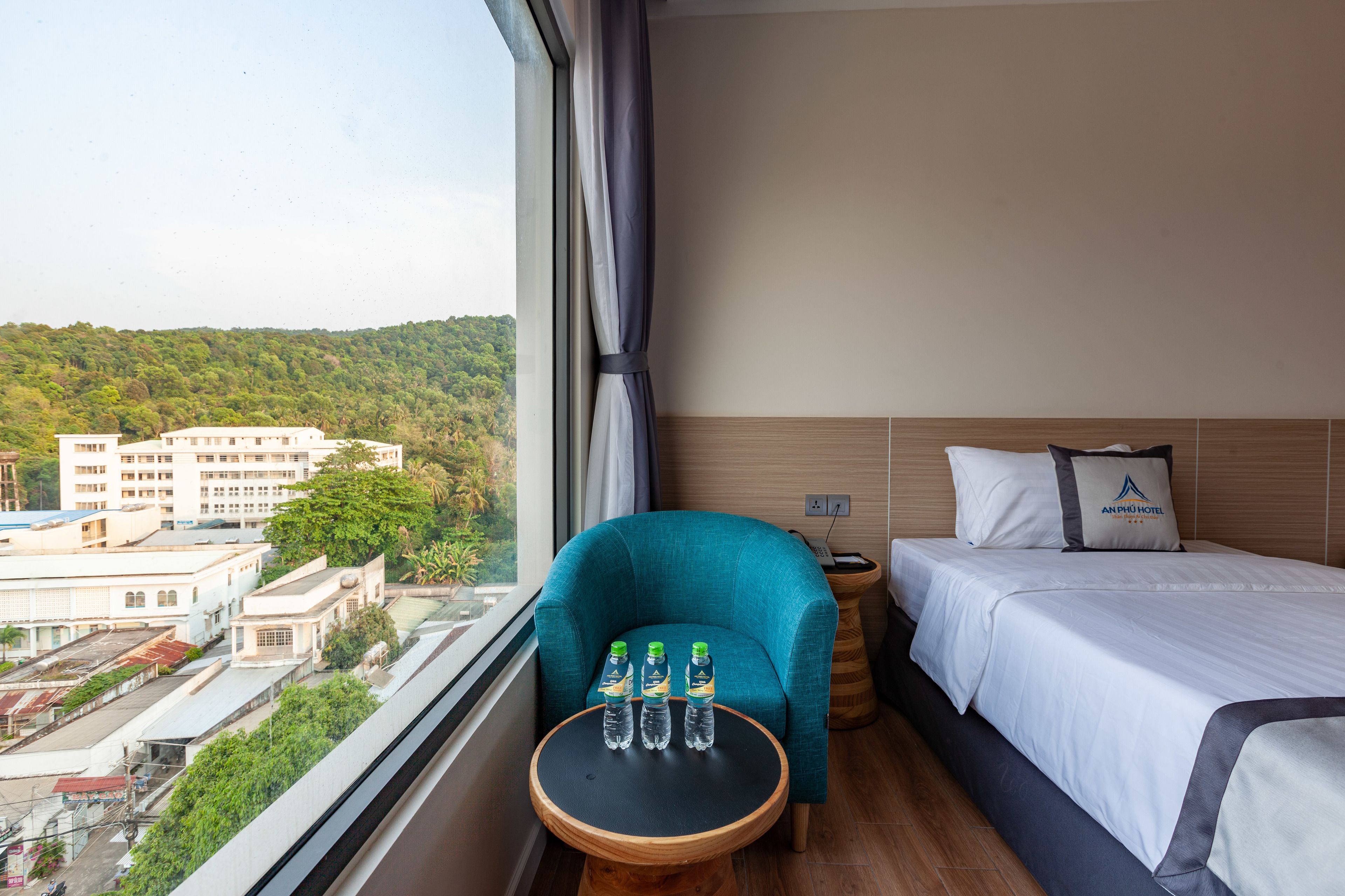 A hotel room with a bed, a blue armchair, a small wooden table with water bottles, and a view of buildings and greenery outside.