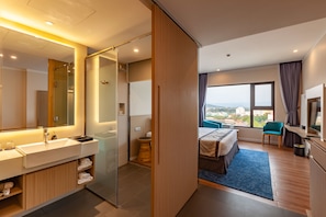 Ein modernes Hotelzimmer mit einem großen Spiegel, einem Badezimmer mit Duschkabine aus Glas, einem Bett mit blauem Teppich, einem Schreibtisch und Blick auf die Stadt.