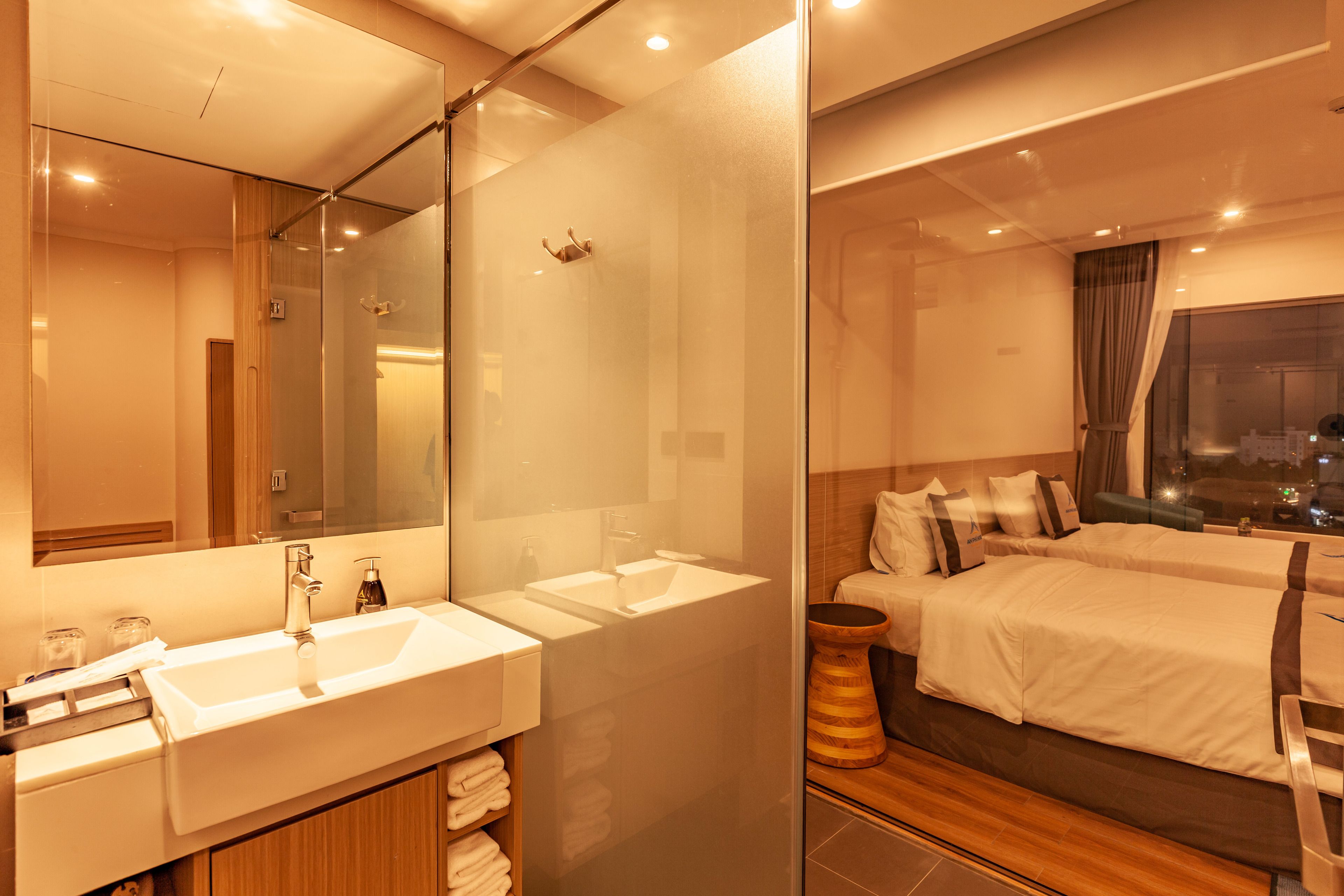 A modern hotel room with a large mirror, two sinks, a wooden vanity, and a bed with a view of the city at night.