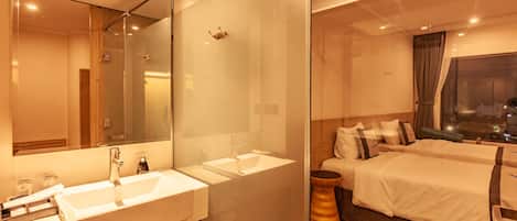 A modern hotel room with a large mirror, two sinks, a wooden vanity, and a bed with a view of the city at night.