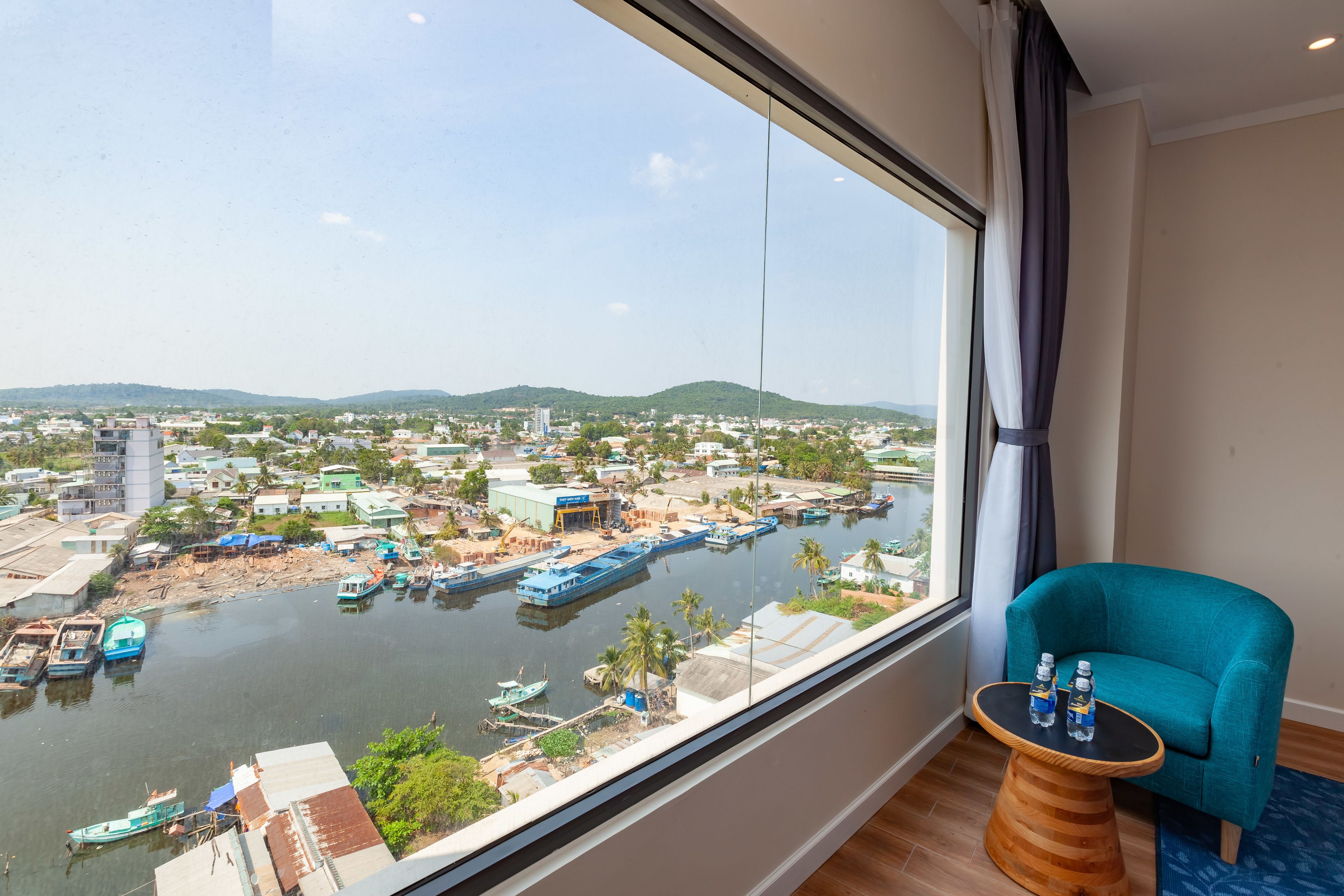 A room with a large window overlooking a river with boats, a teal armchair, and a small wooden table with bottles.