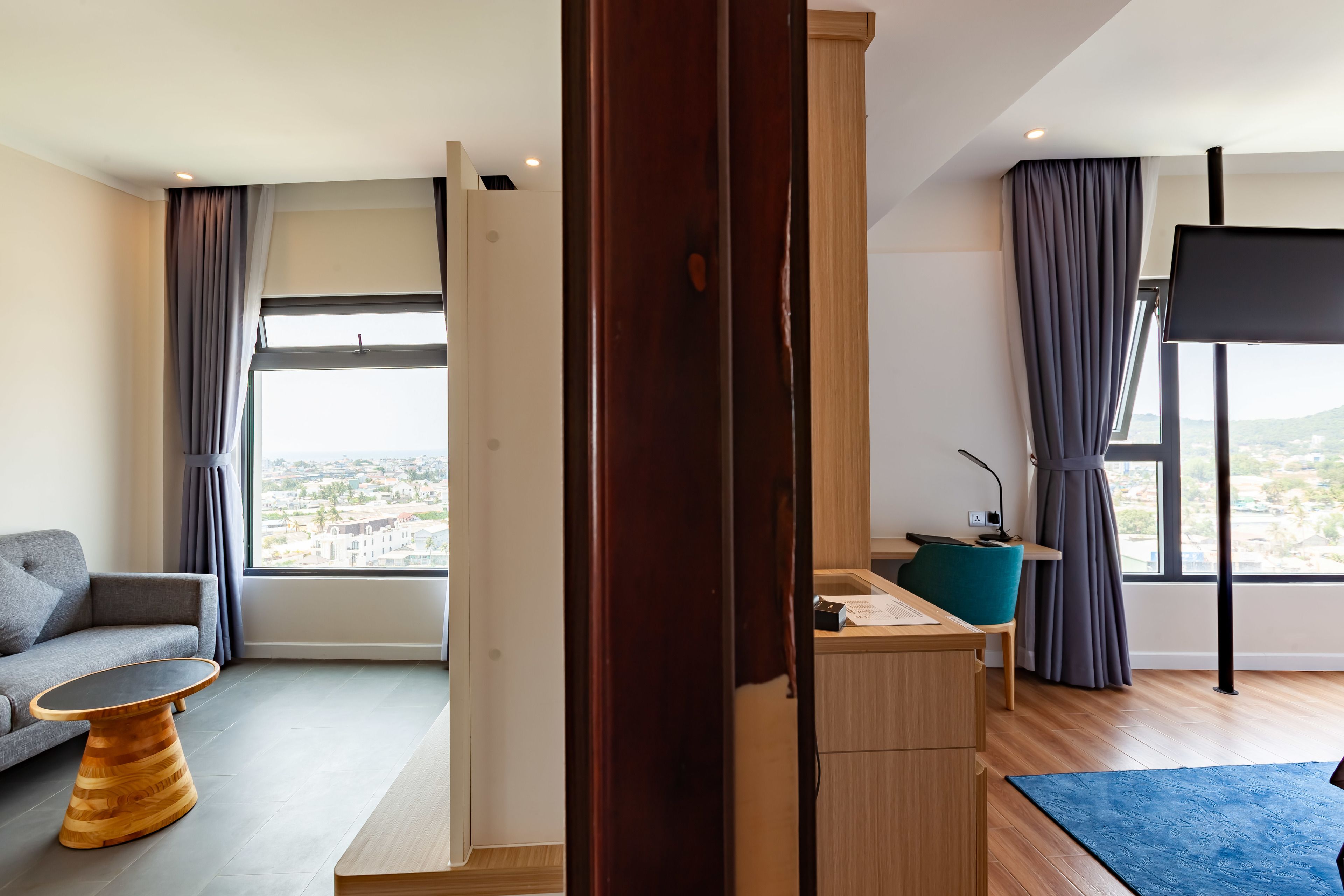 A modern hotel room with a sofa, a wooden coffee table, a desk with a chair, and a large window offering a city view.