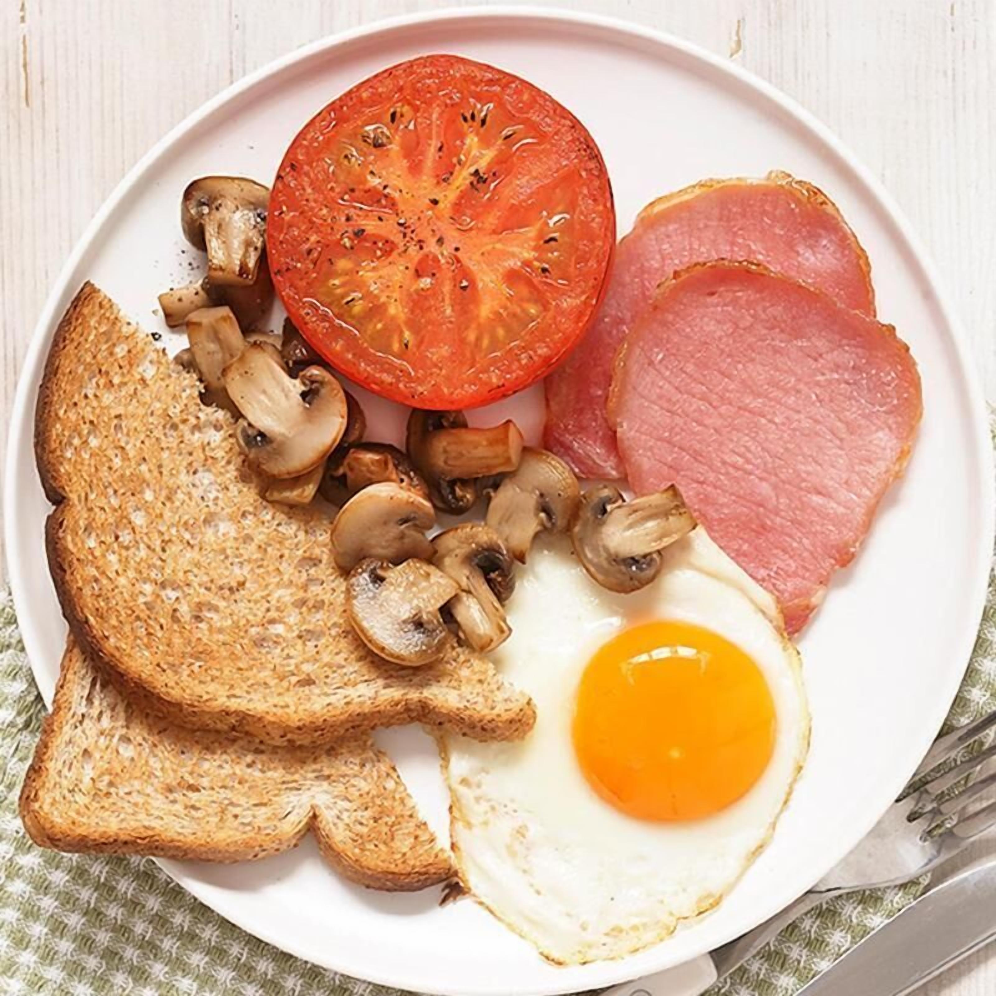 Un plato con una rodaja de tomate, un huevo frito, una loncha de jamón, una tostada y varios hongos.