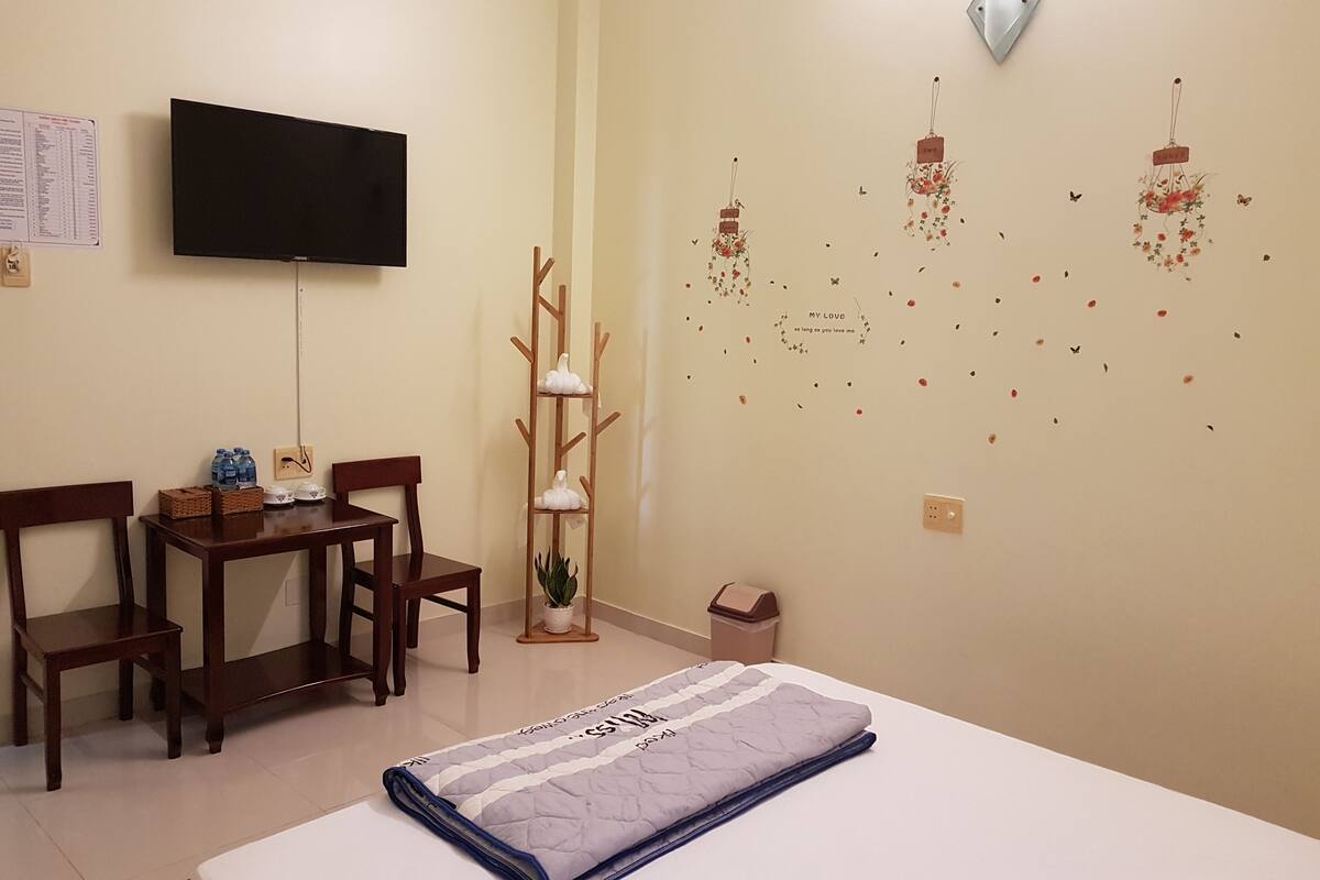 A hotel room with a bed, a desk with chairs, a television, a wall-mounted plant, and decorative wall art.