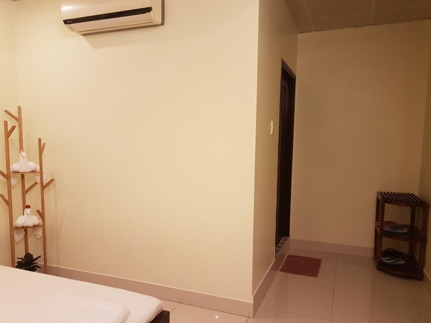 A hotel room with a bed, an air conditioning unit, a wooden shelf with towels, and a shoe rack.