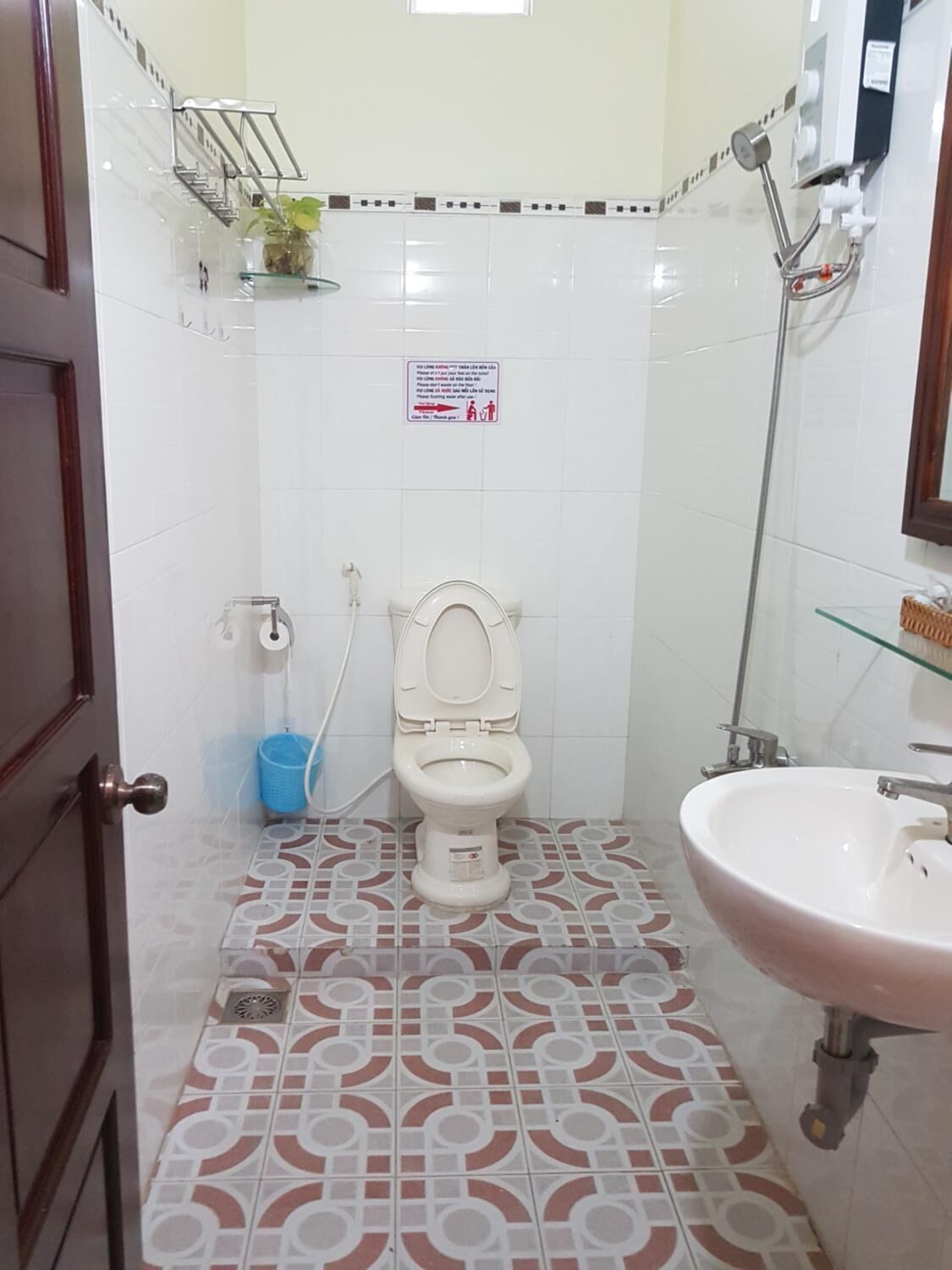 A bathroom with a toilet, sink, and shower. There is a sign on the wall and a plant on a shelf.