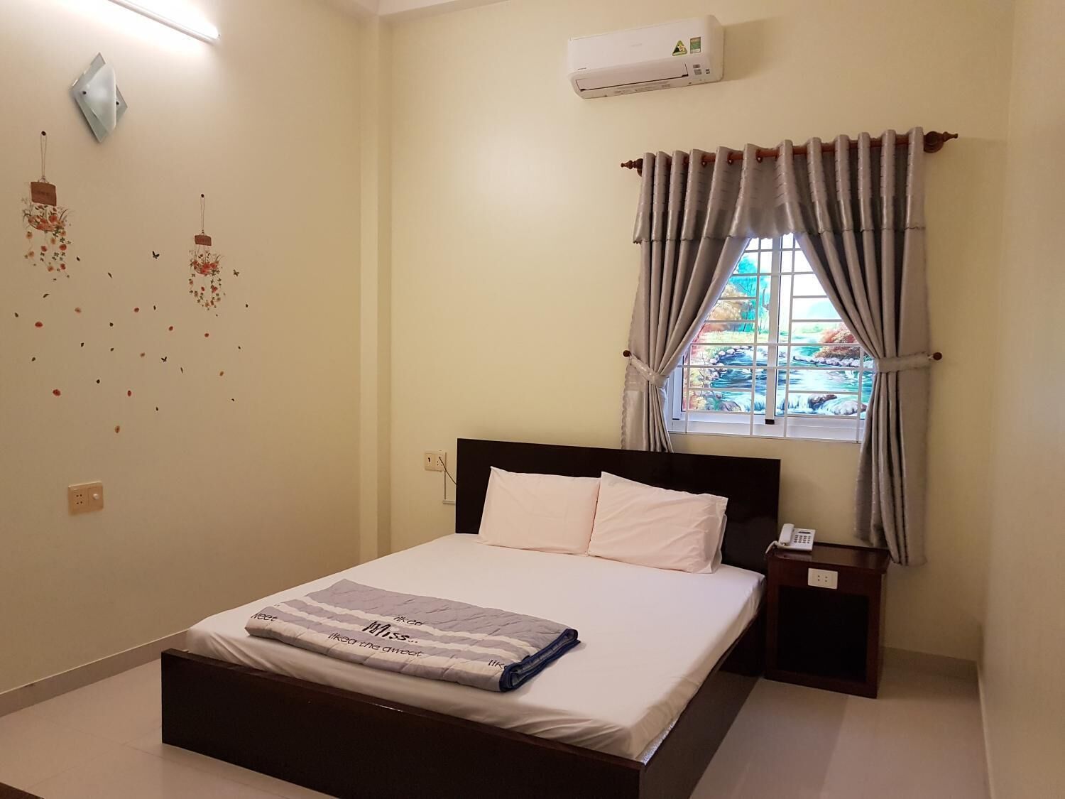 Una cama bien tendida con edredón blanco y almohadas, una mesita de noche con un teléfono, una ventana con cortinas y un equipo de aire acondicionado en la pared.