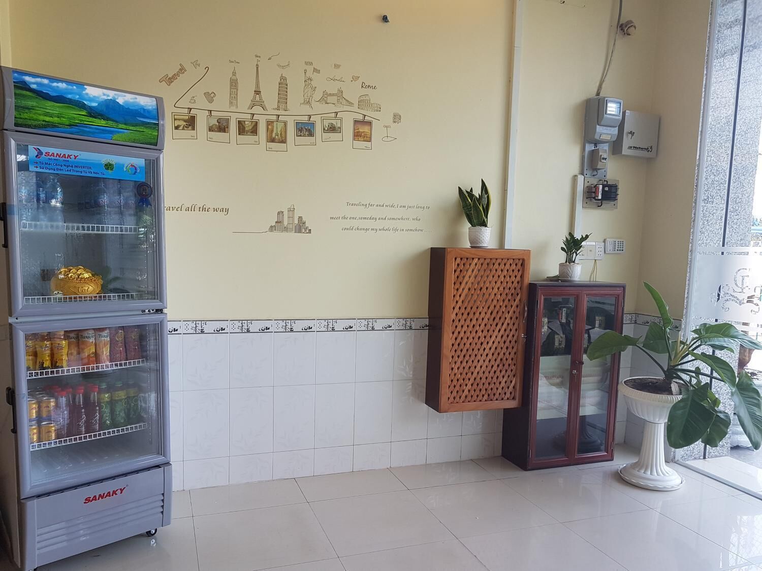 Un espace d’accueil comprenant un réfrigérateur rempli de boissons, un mur décoré de photos de voyages et d’une citation, ainsi qu’une plante en pot.